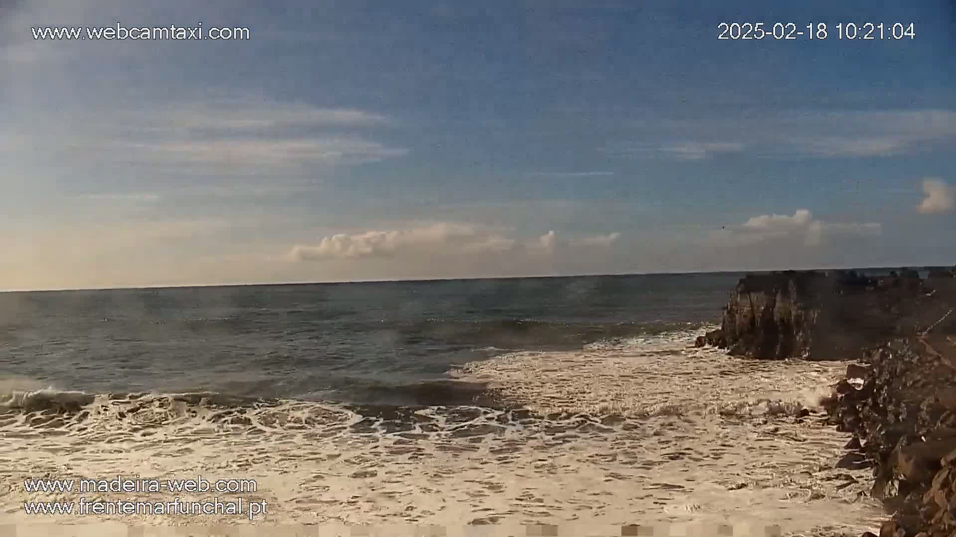 A partly cloudy sky overlooks a rocky coastline where waves are crashing against the shore.