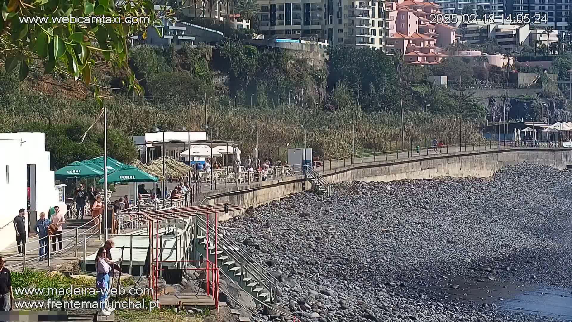 A sunny outdoor scene shows a rocky shoreline with a paved walkway, several outdoor restaurants, and people walking along the path.