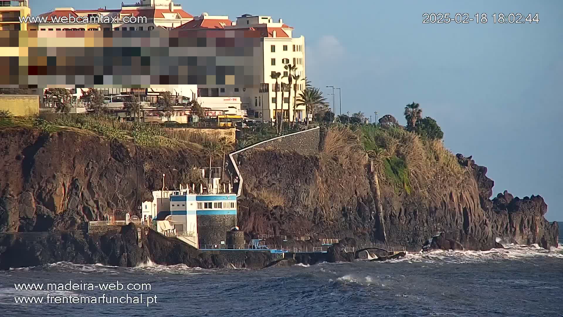 A seaside cliff with a building on it, overlooking a wavy ocean under a clear sky.