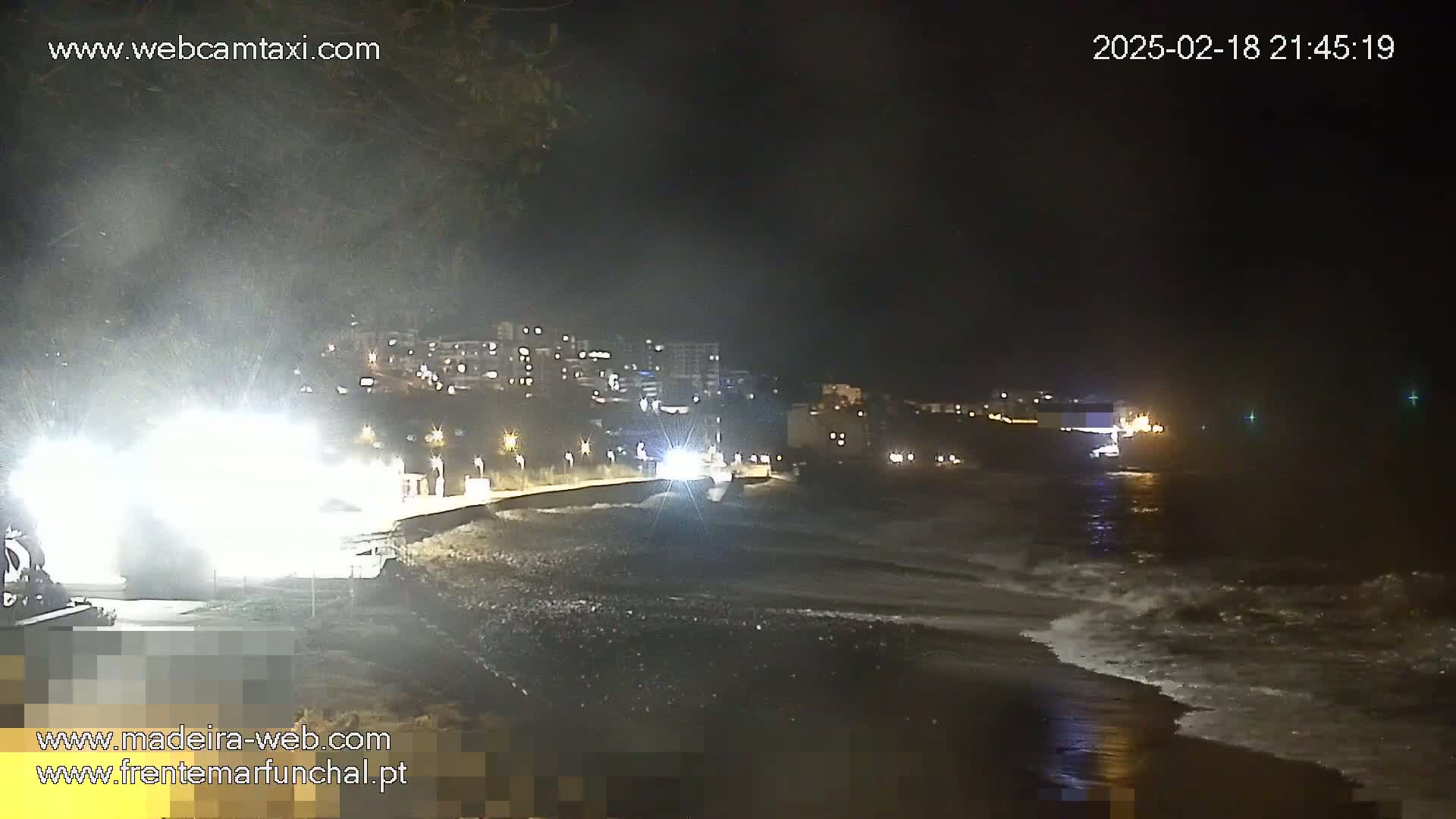At night, moderately rough waves lap a dark beach next to a dimly lit town under a hazy sky.