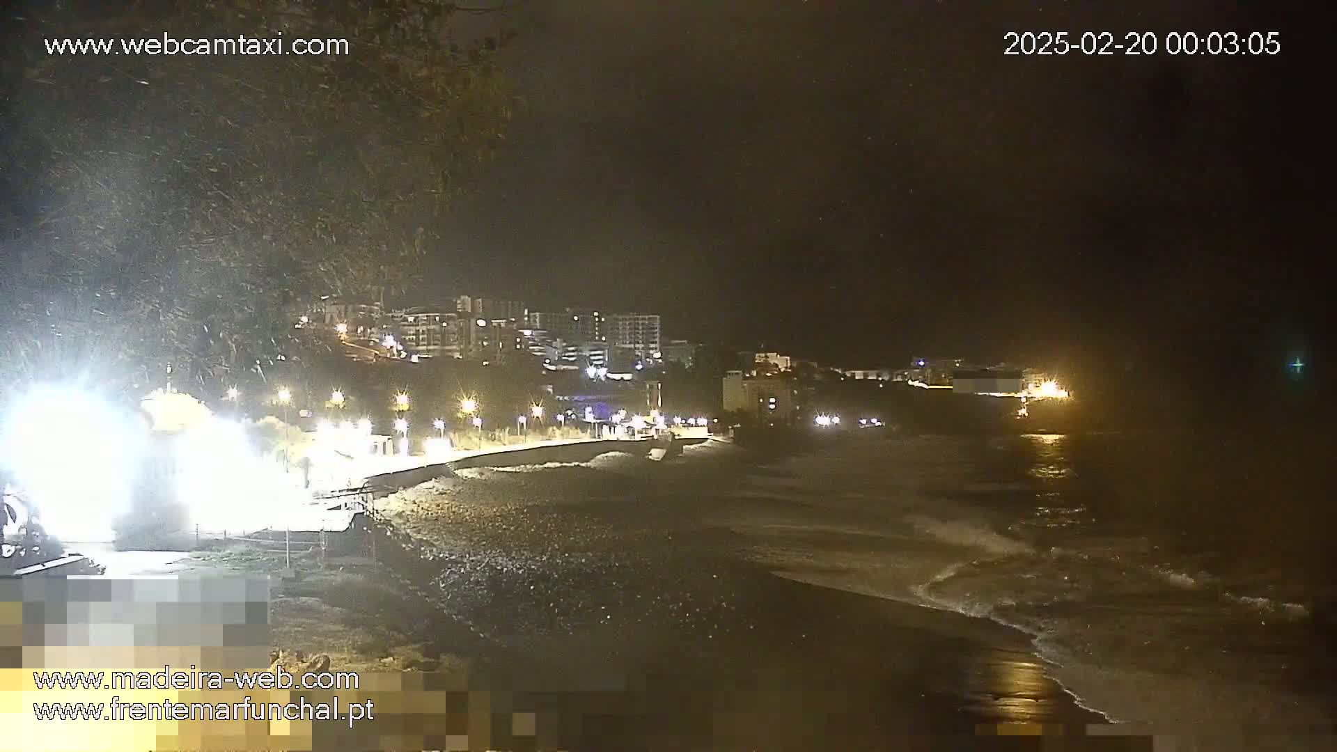 A nighttime coastal scene shows a dark, wavy ocean lapping against a pebble beach, with buildings and lights visible in the distance under a dark, possibly stormy sky.