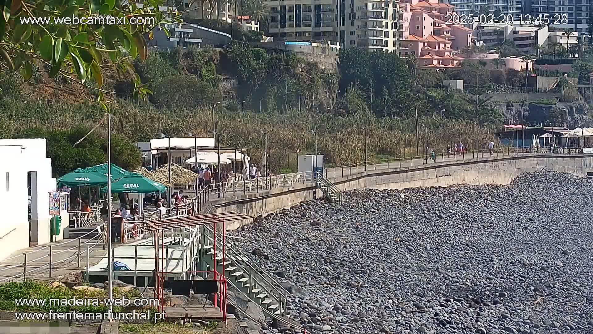 A rocky beach with a paved walkway lined with outdoor seating and buildings, under a sunny sky.