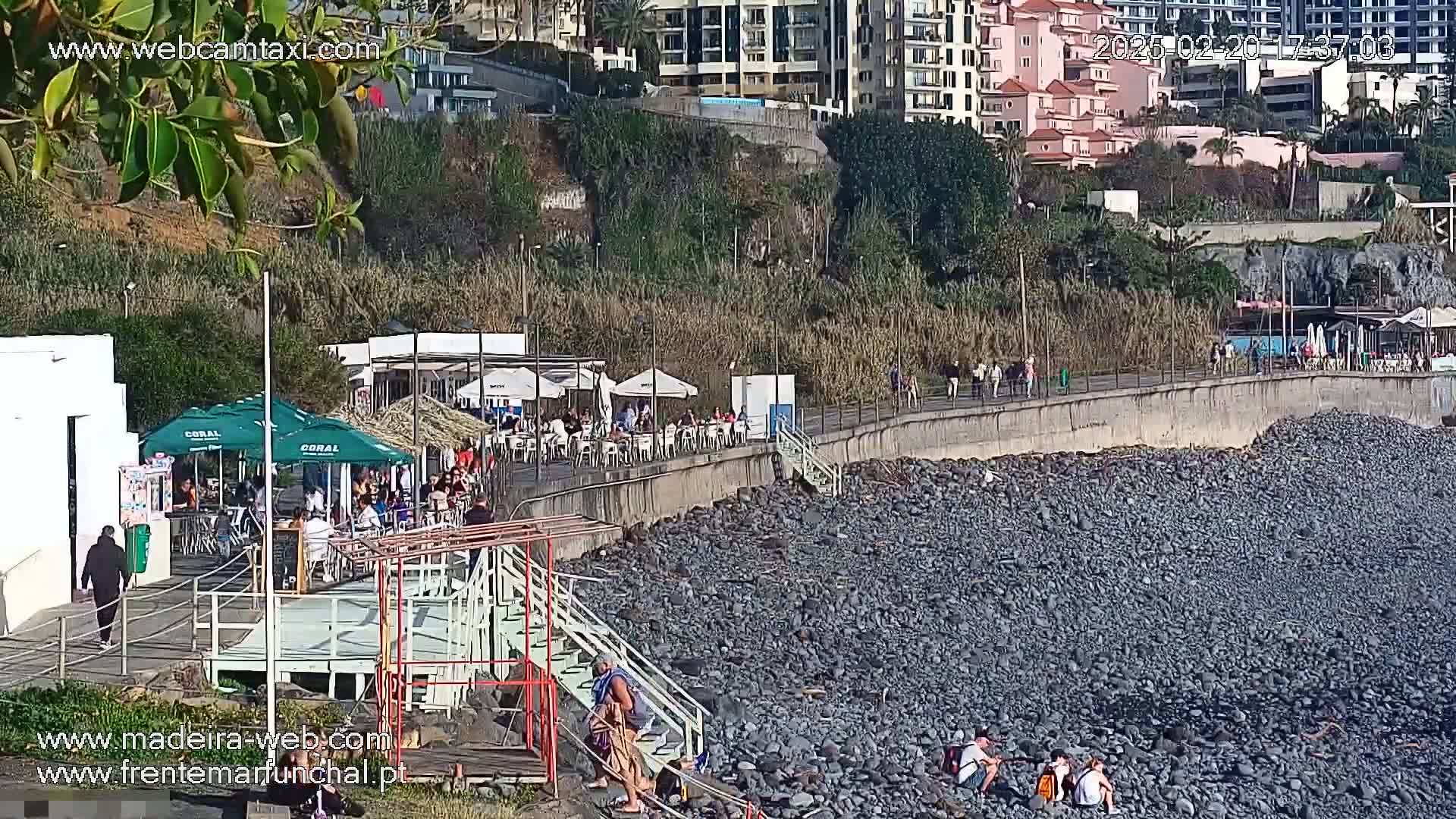 A seaside restaurant with outdoor seating overlooks a rocky beach on a sunny day.