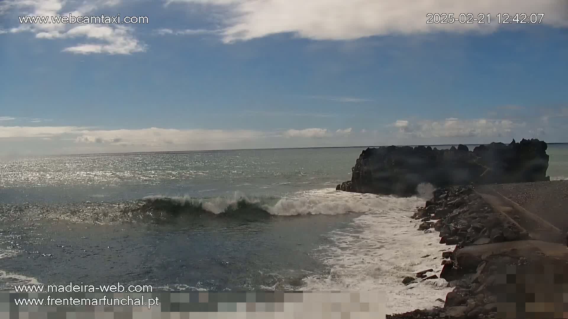 Ocean waves crash against a rocky shoreline under a partly cloudy sky.