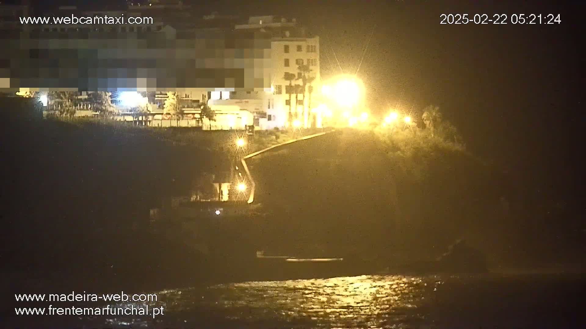 A nighttime coastal scene shows a building lit by streetlights overlooking a dark body of water under a hazy sky.