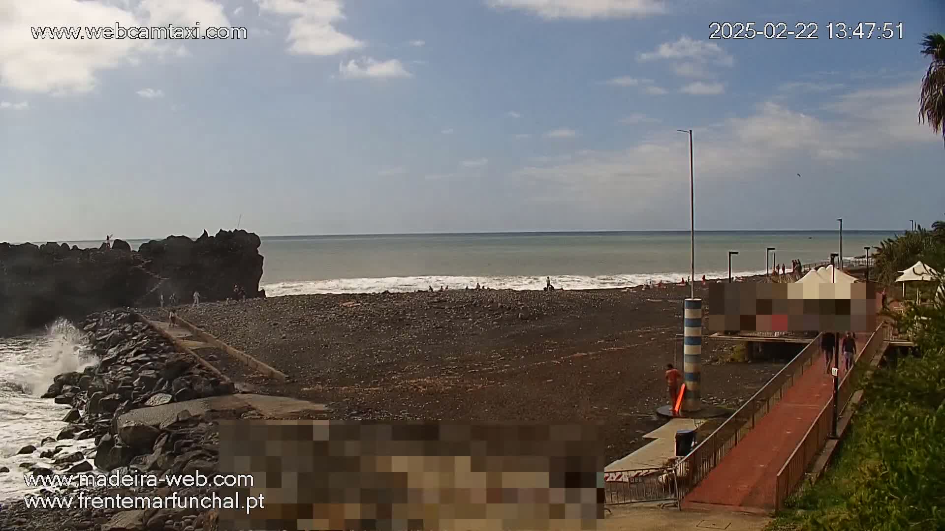 A dark-sand beach with a few people on it and some waves crashing against a rocky jetty, under a partly cloudy sky.