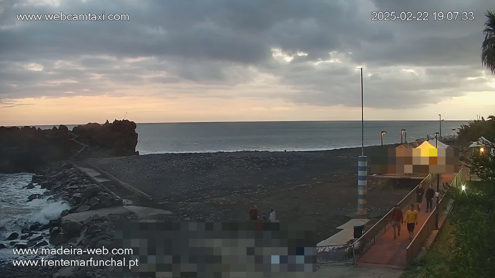 A dark, rocky shoreline with a few people near a structure under a cloudy sunset sky, with some waves crashing against the shore.