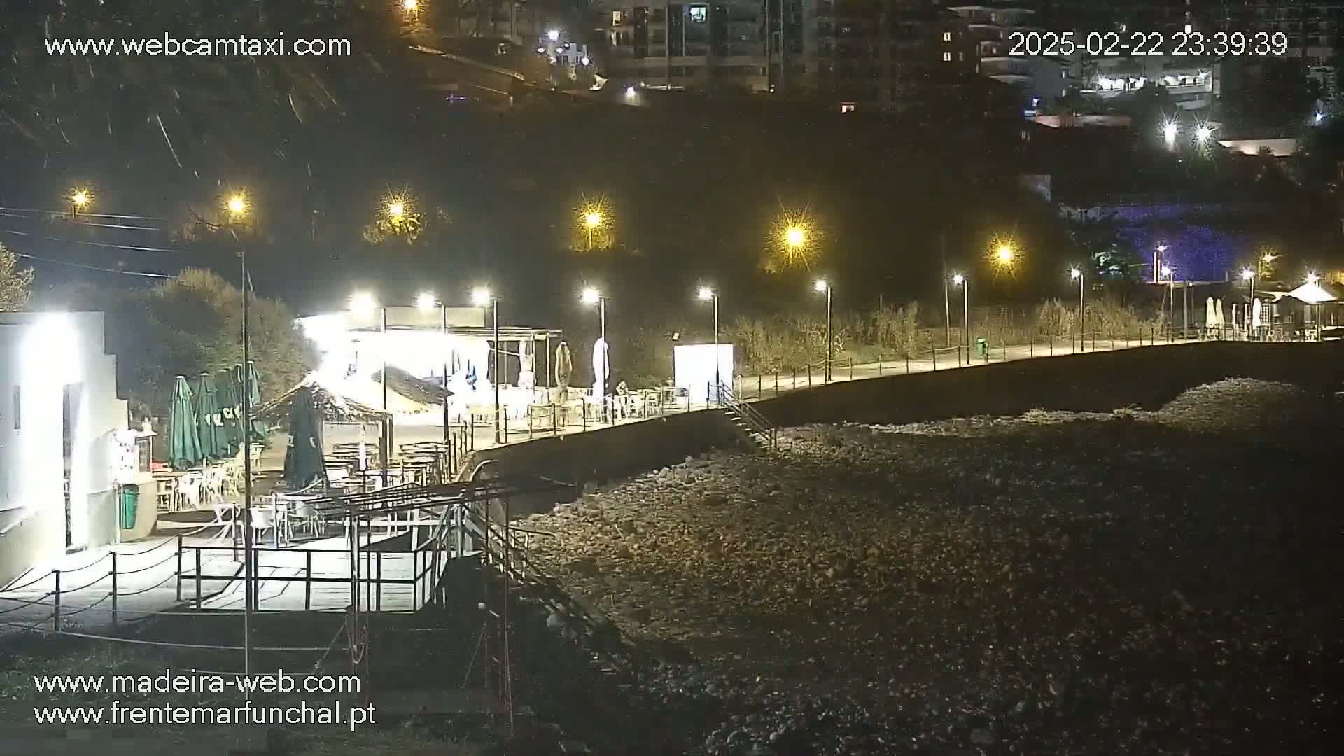 A nighttime view of a shoreline with a boardwalk, outdoor seating, and lights, overlooking a dark, rocky beach.