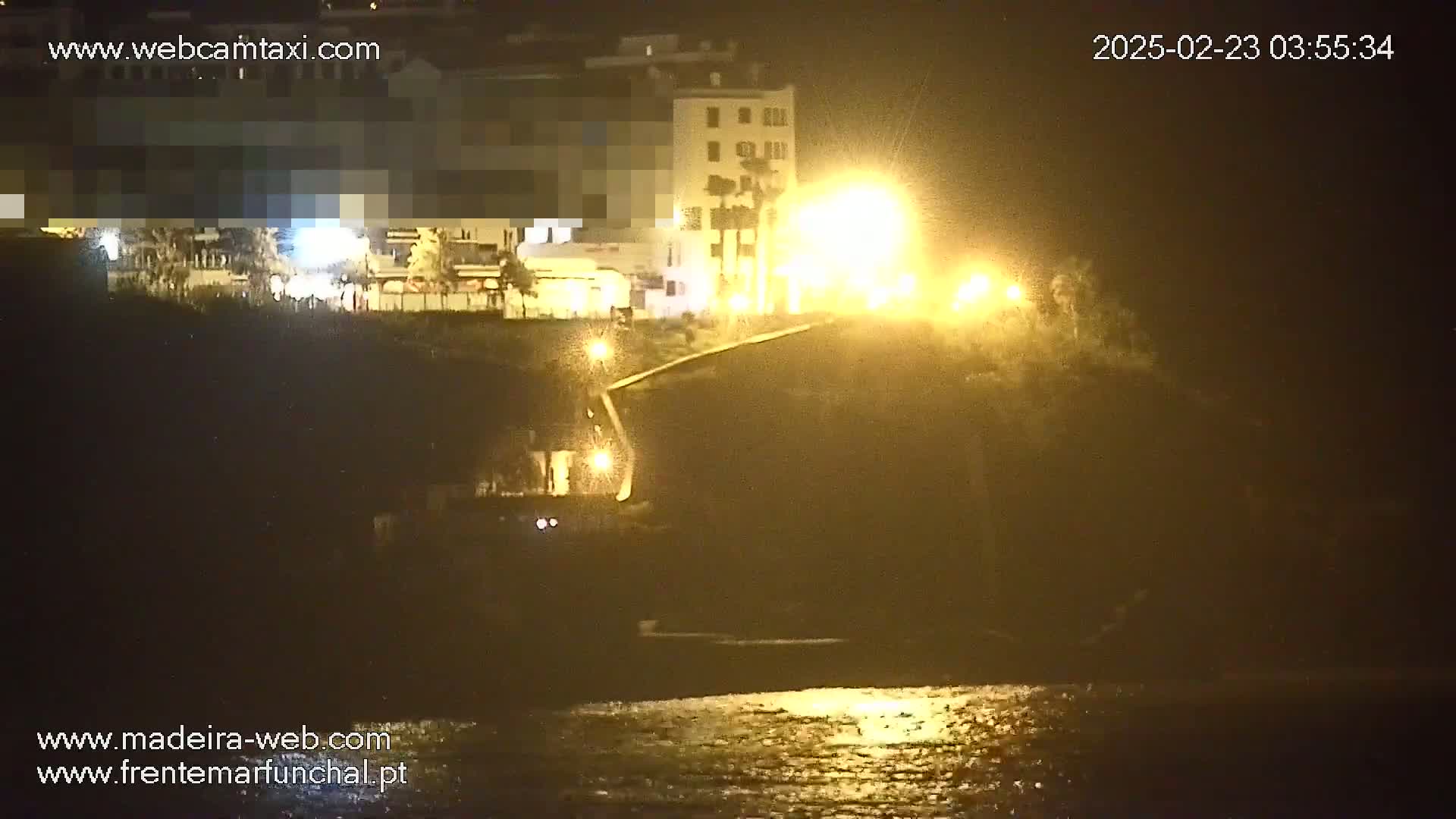 A nighttime coastal scene shows a dark cliff and buildings illuminated by streetlights, with a calm body of water reflecting the lights in the foreground.