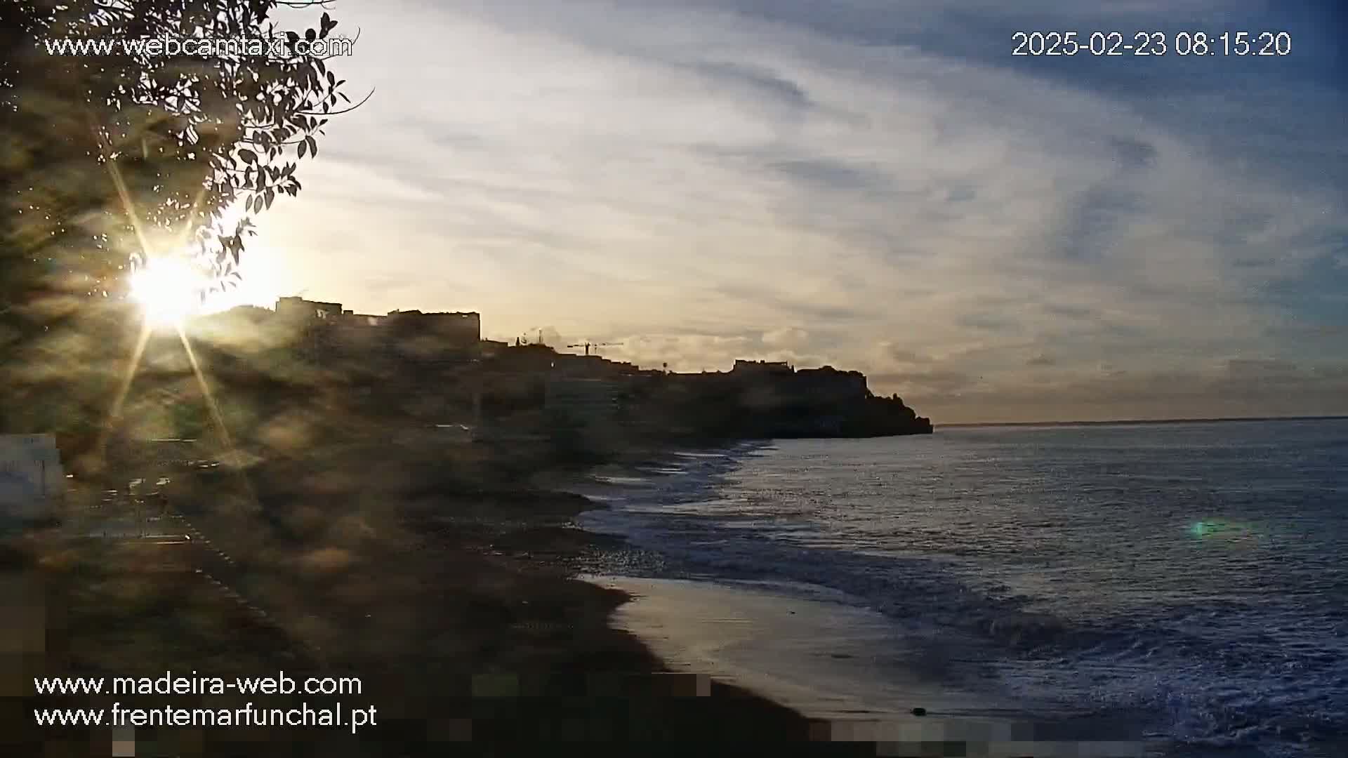 A partly cloudy sunrise illuminates a dark, sandy beach and buildings silhouetted against a calm ocean.
