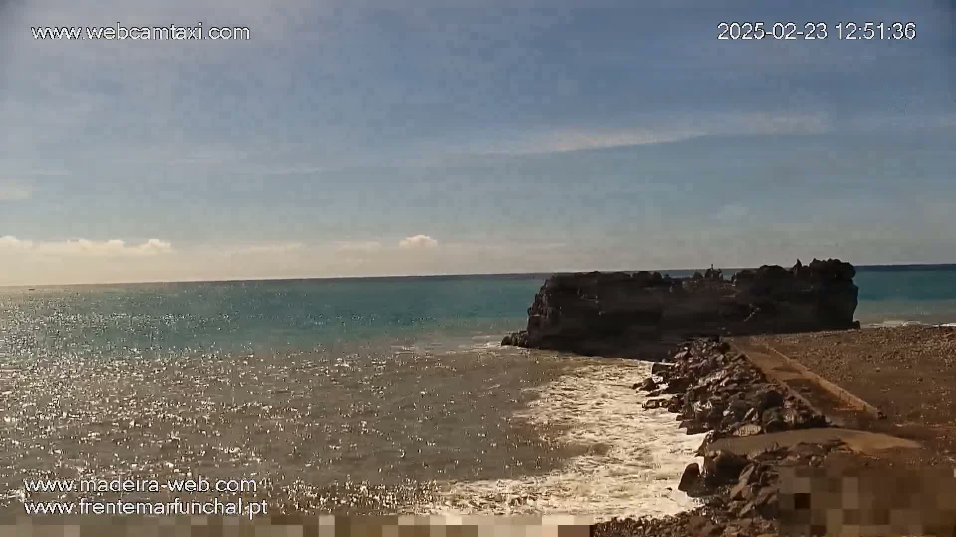 A rocky outcrop juts into a calm, teal ocean under a mostly clear sky.