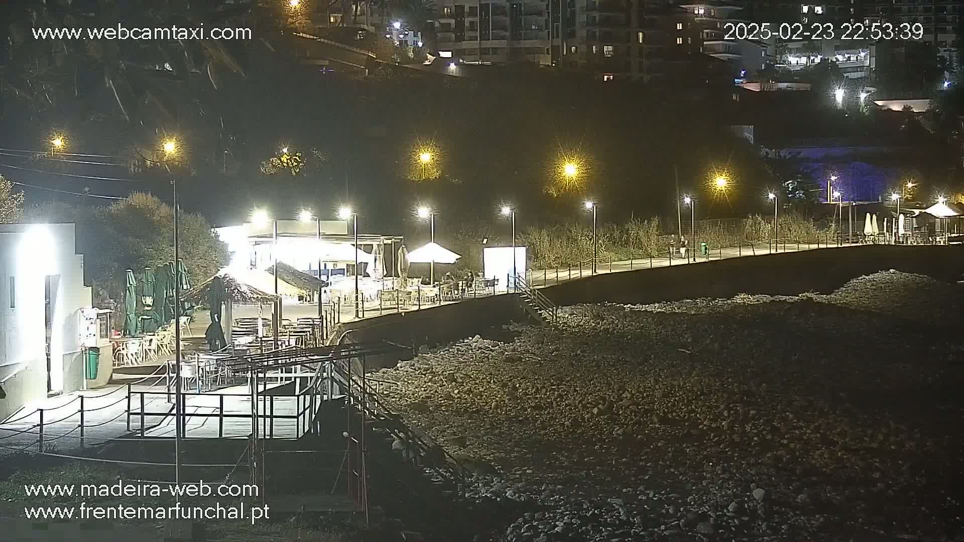 A nighttime view of a rocky beach with outdoor seating areas and lights illuminating a boardwalk next to a hillside.