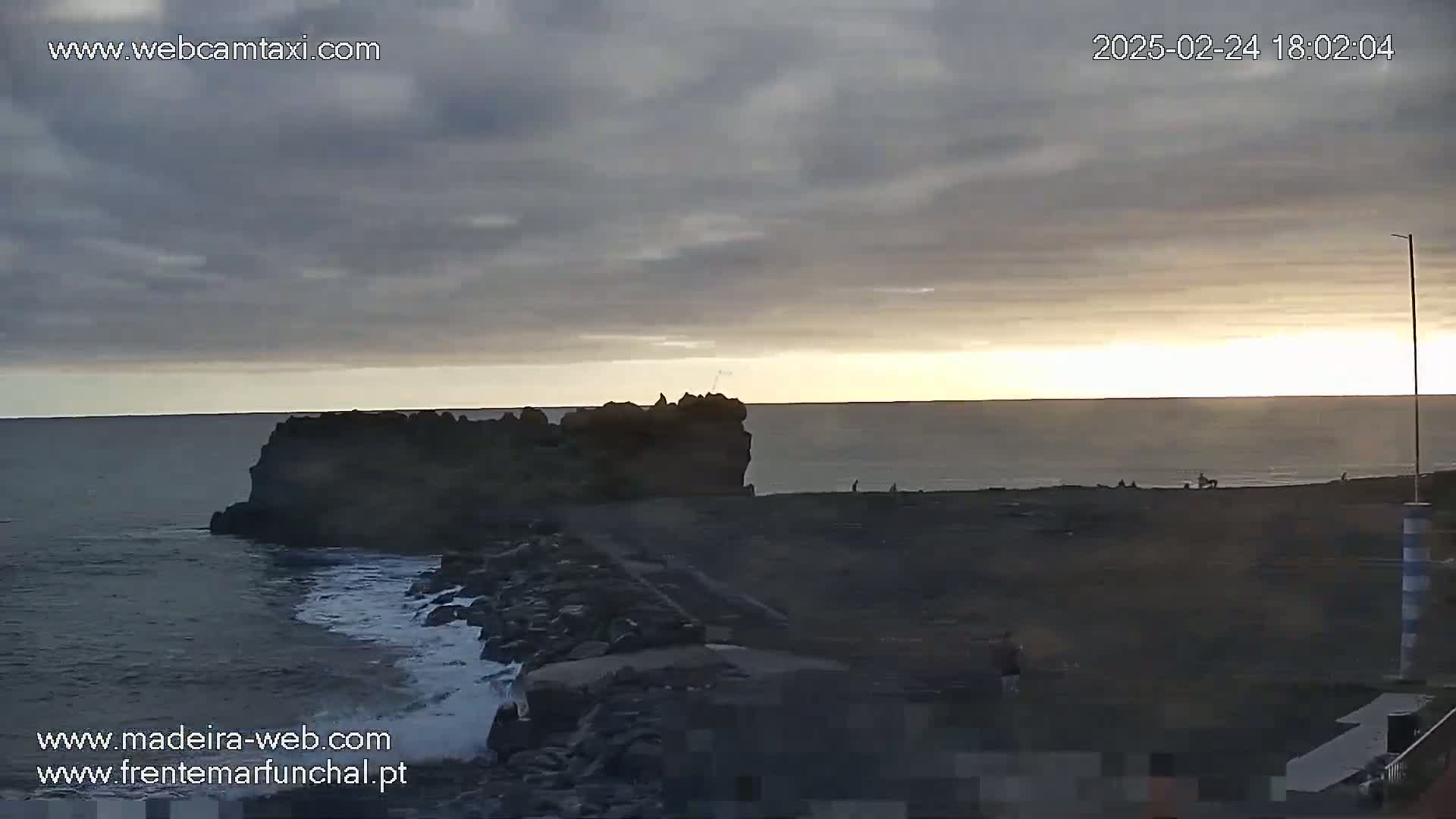 A rocky coastline with a dark, overcast sky shows a sunset over a calm ocean with a few people visible on the shore.