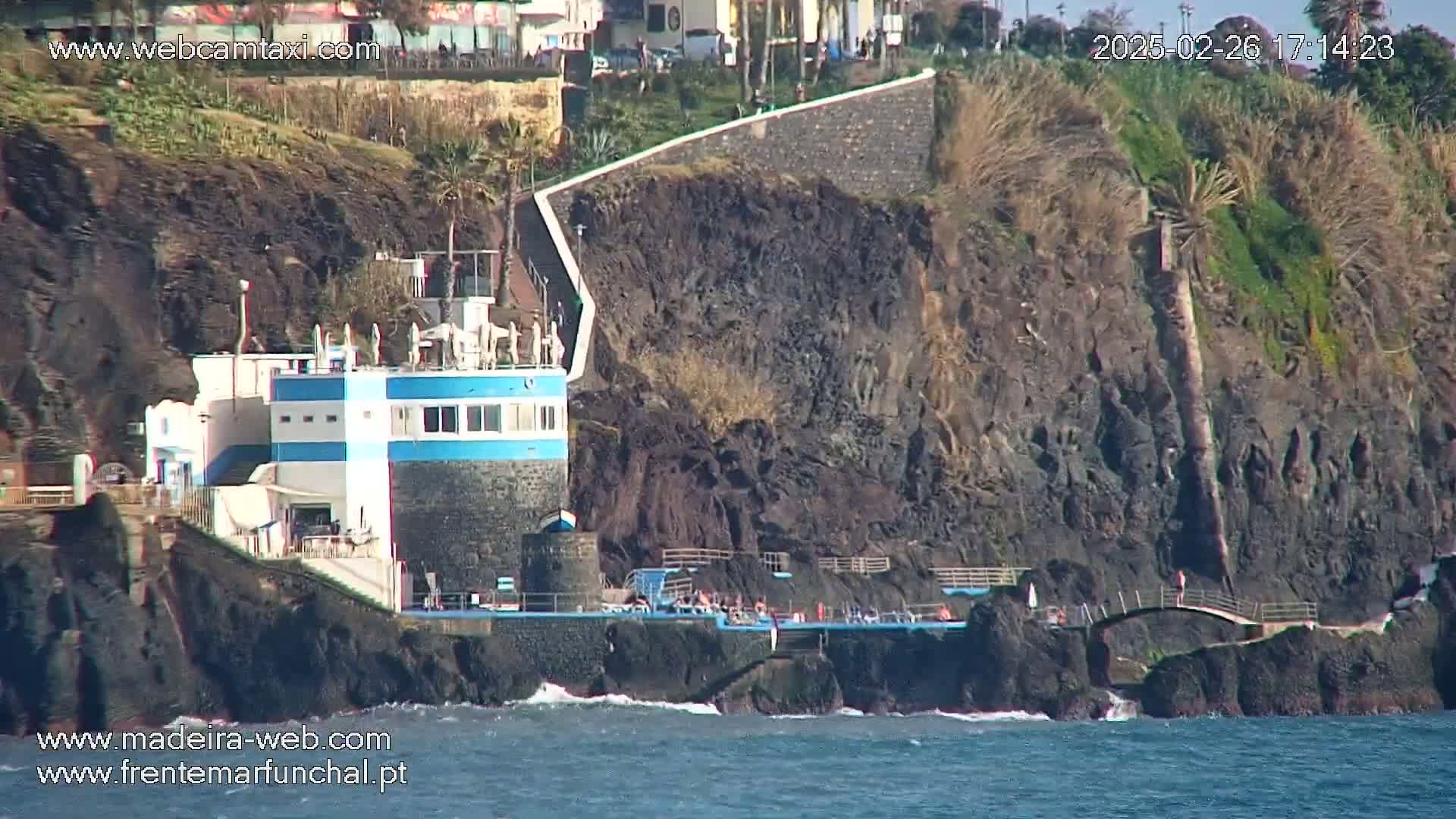 A seaside building with blue trim sits on a rocky cliff overlooking a dark-blue ocean with small waves under sunny skies.