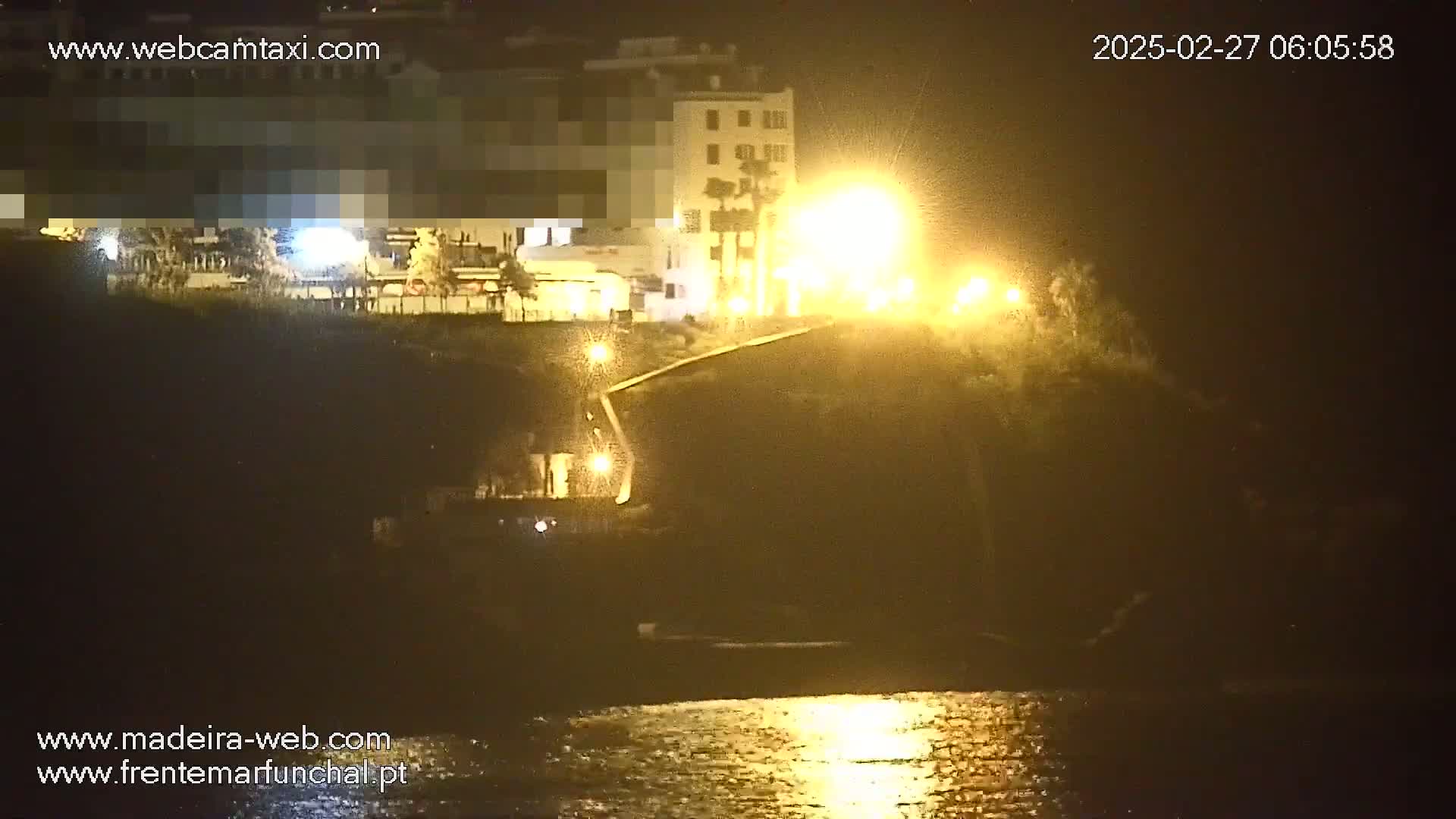 A nighttime, brightly lit waterfront scene shows a building perched on a cliff overlooking calm water.