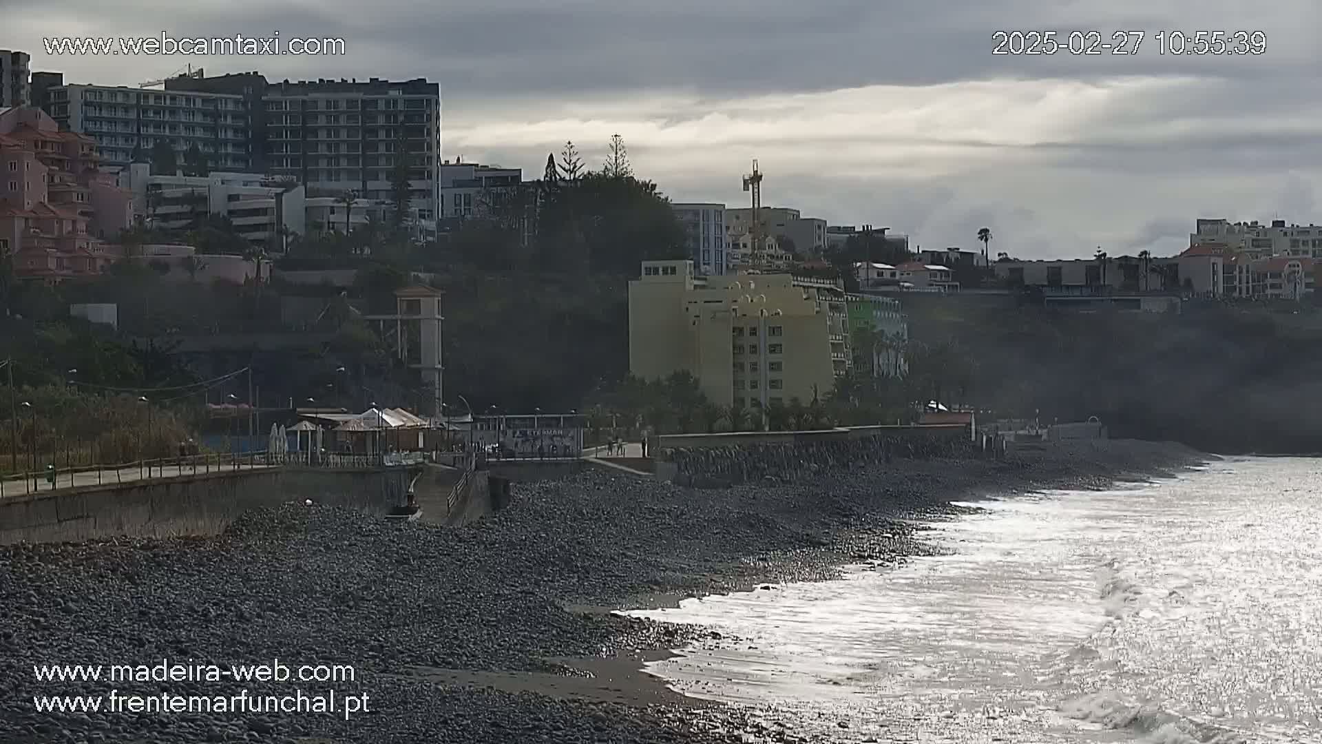 A rocky beach with gentle waves laps the shore under an overcast sky, with buildings and hotels rising on a hillside behind it.