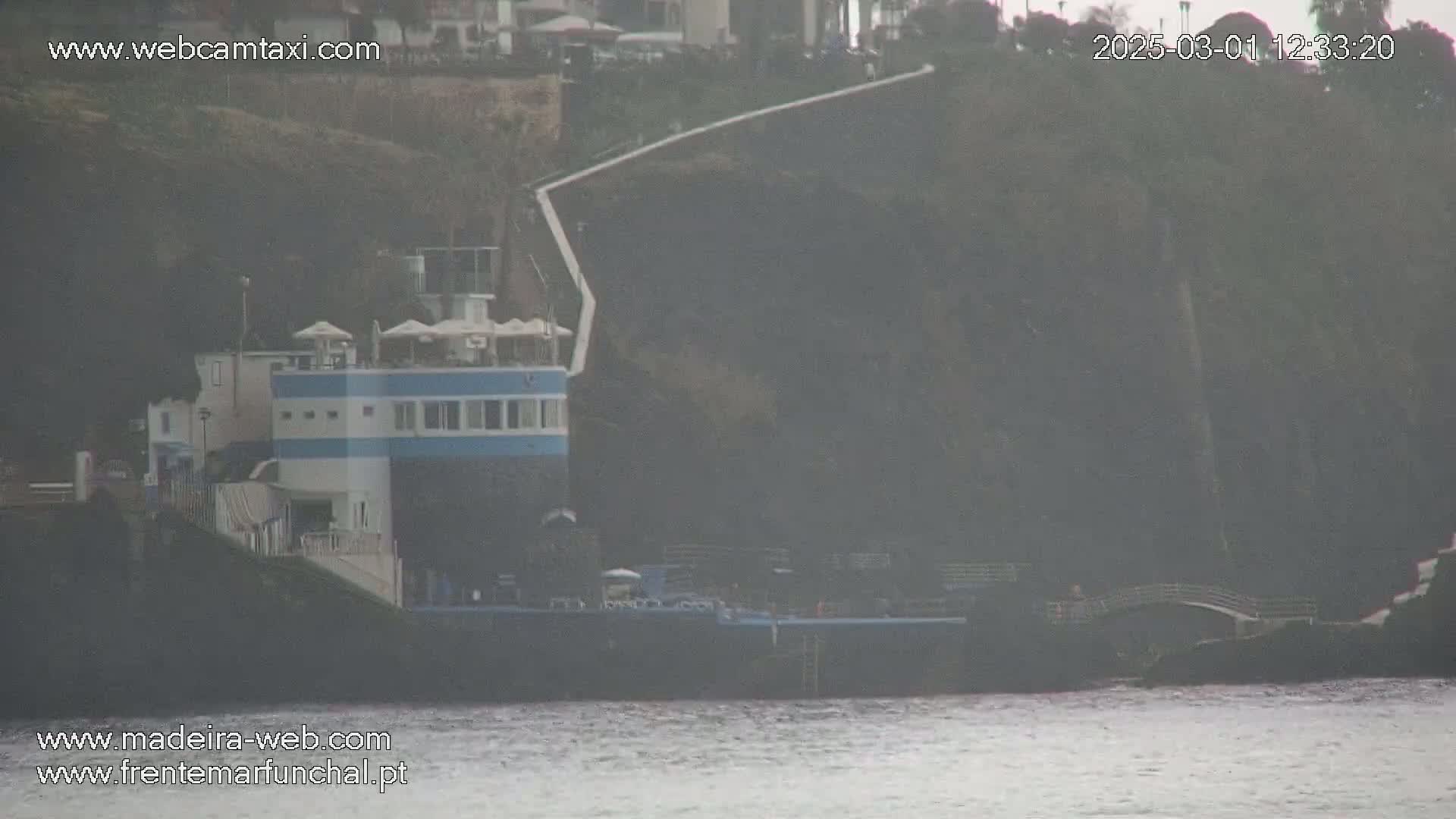 A hazy view shows a blue and white building next to a body of water, with a dark cliff behind it.