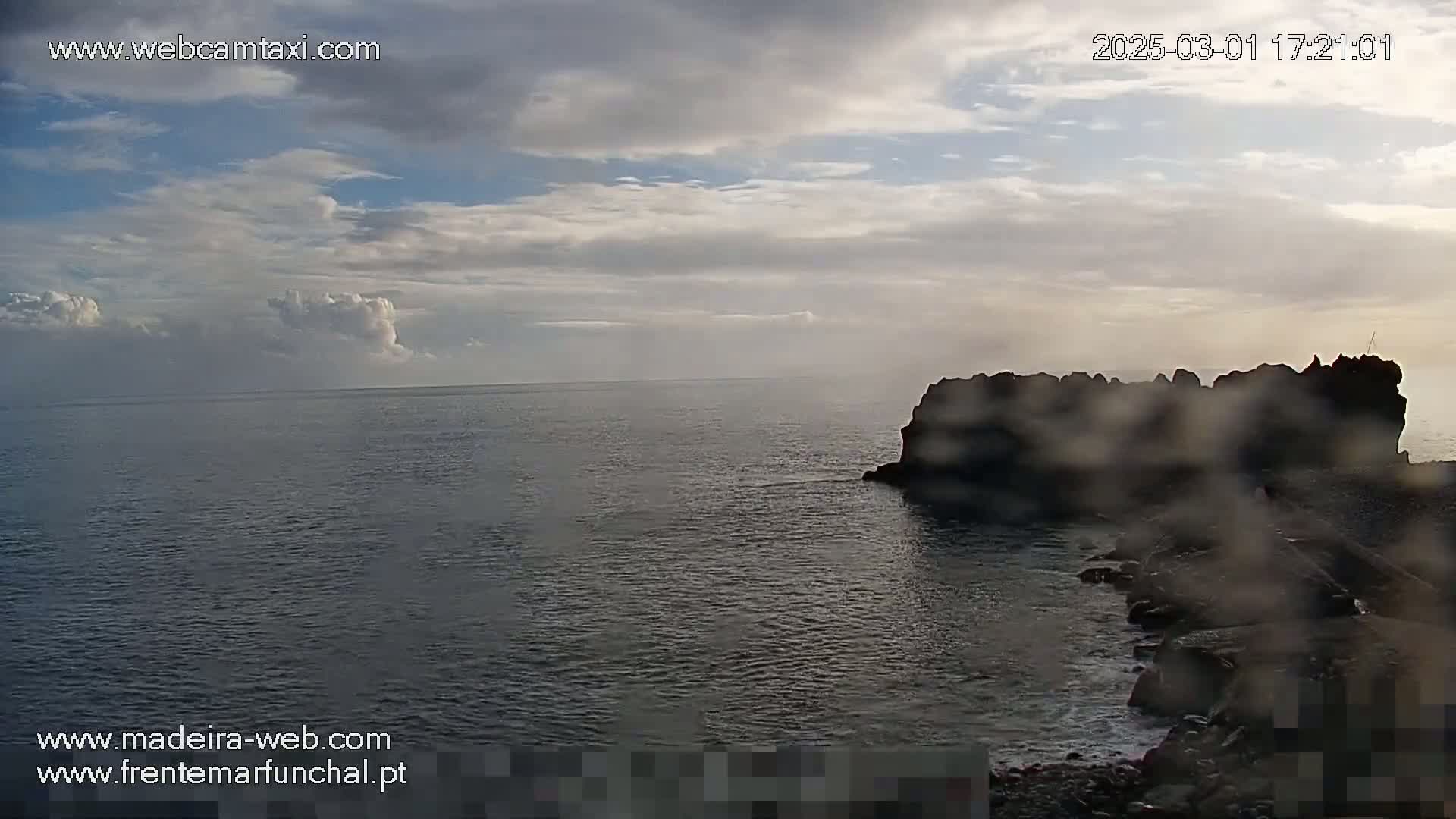 A partly cloudy sky overlooks a dark body of water with a rocky outcrop in the mid-ground.