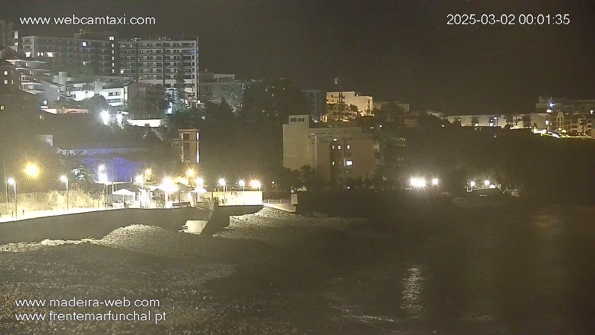 A nighttime view of a rocky shoreline with buildings illuminated on a hill above, under a dark, possibly cloudy sky.