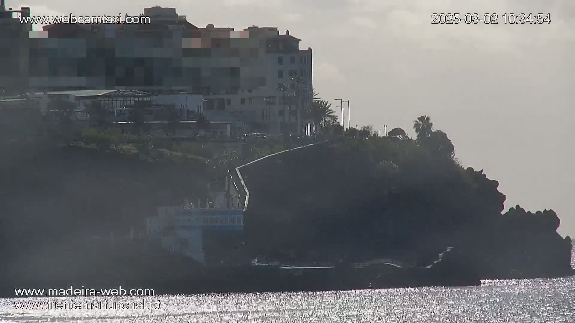 A multi-story building sits atop a dark cliff overlooking a calm, glittering body of water under an overcast sky.