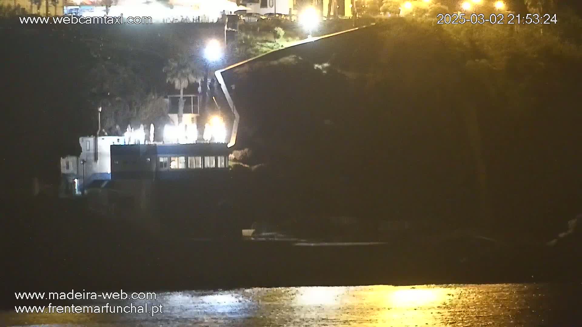 A dark building sits on a waterfront at night, illuminated by artificial lights, overlooking calm water.