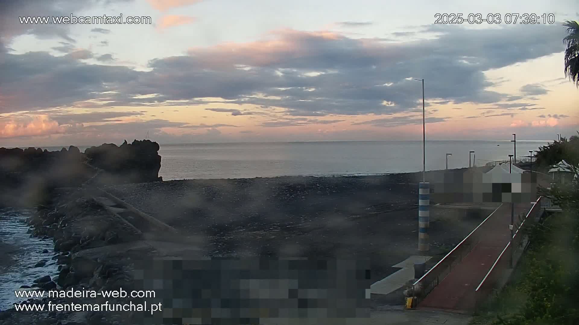 A dark, rocky shoreline meets a calm ocean under a cloudy, pinkish-hued sunrise.