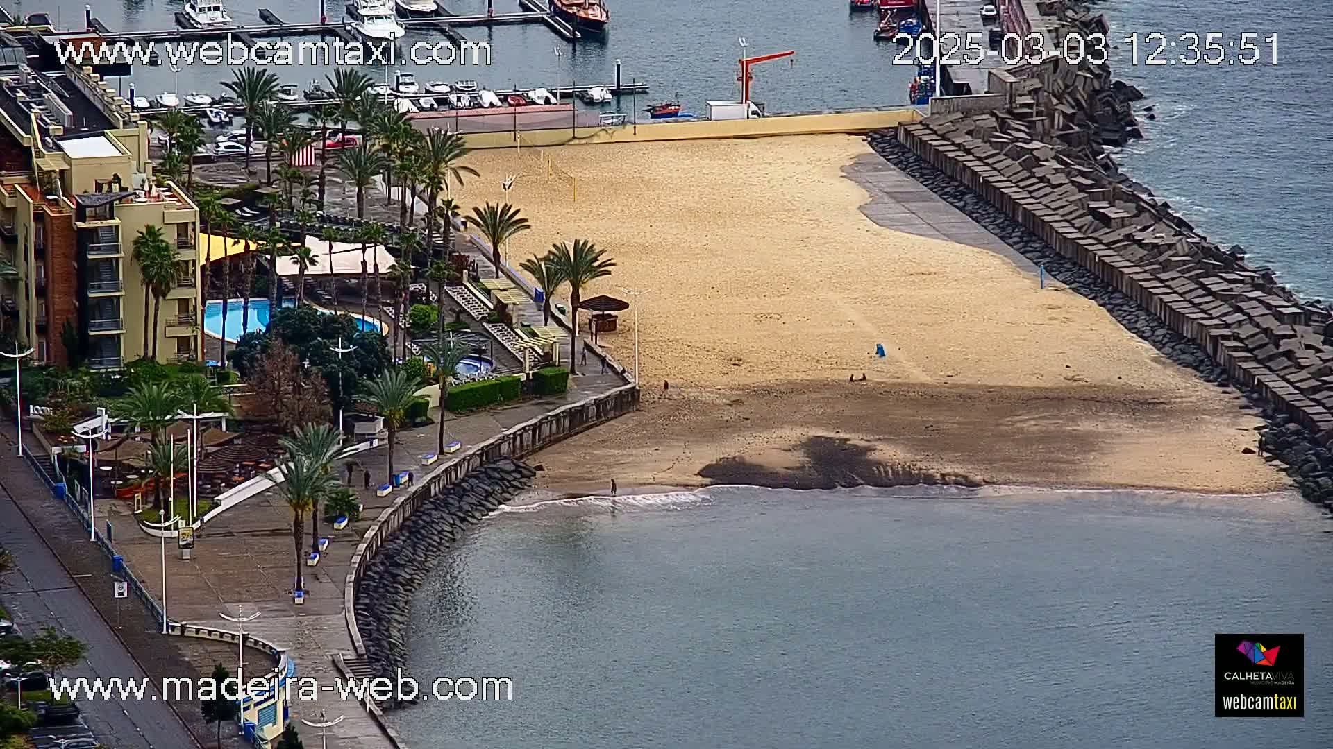 An aerial view shows a sandy beach bordered by a stone wall, a hotel with a pool, palm trees, and calm ocean water.