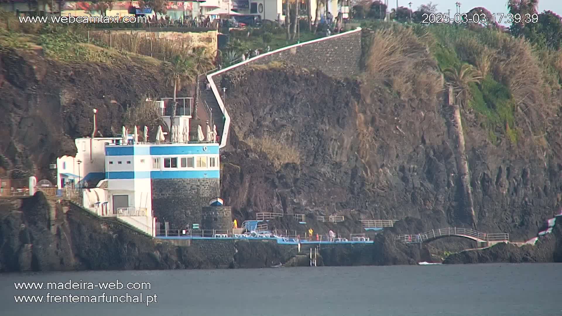 A small, blue-and-white building sits on a rocky cliff overlooking calm water, with a walkway leading up the cliffside.