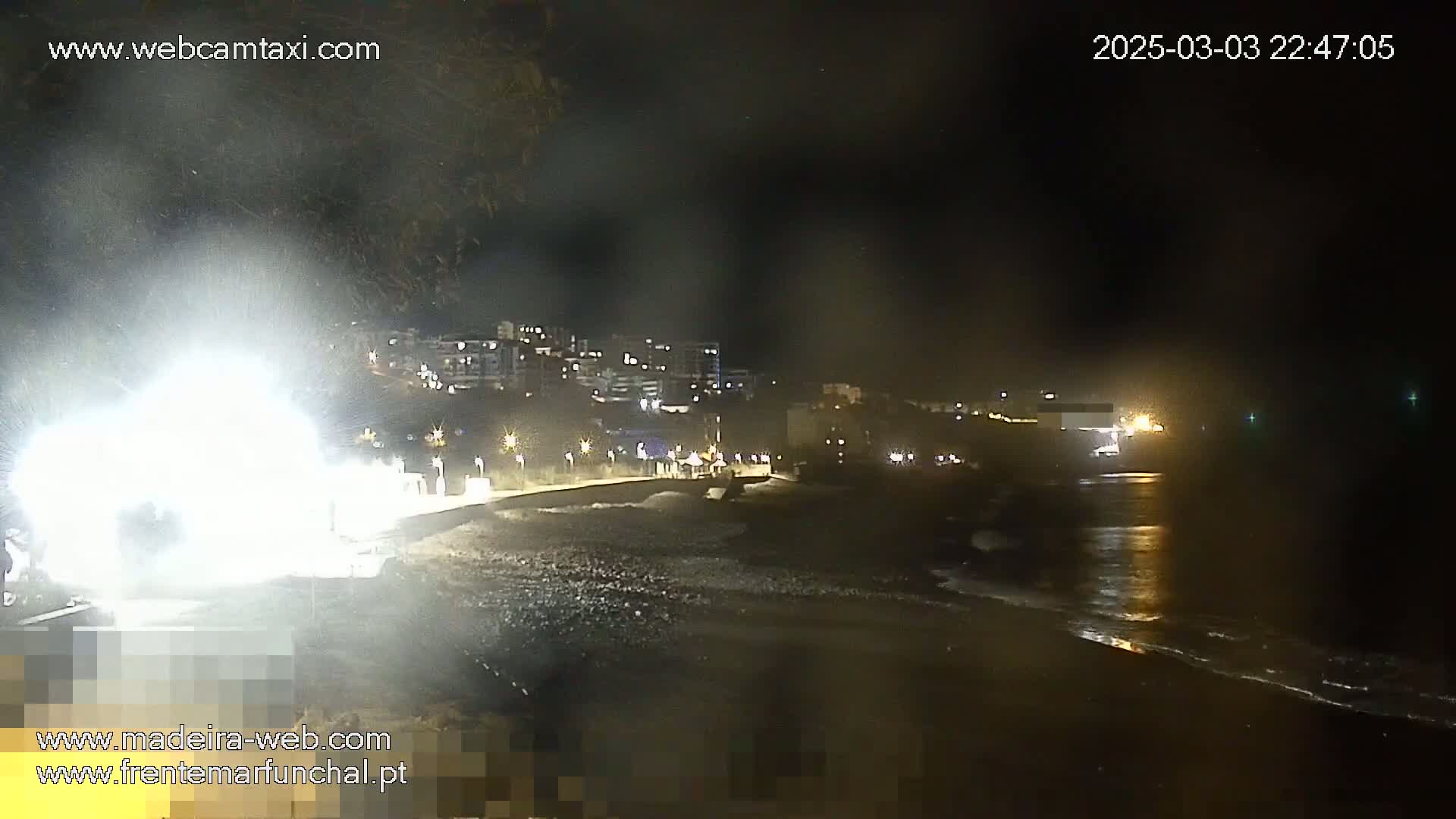 A nighttime view of a dark beach with gently lapping waves, illuminated buildings in the background, and a bright light source obscuring the foreground.