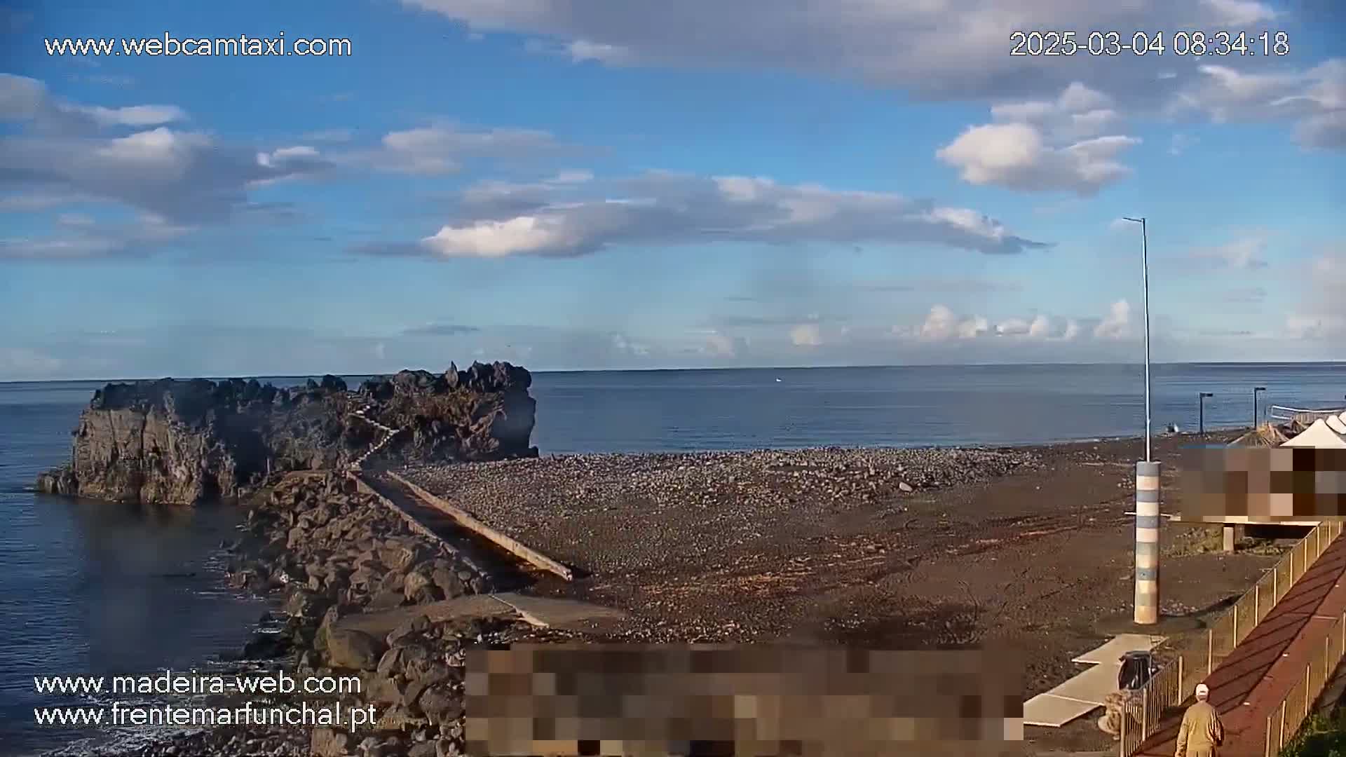 A rocky shoreline with a pebble beach stretches to a calm ocean under a partly cloudy sky.