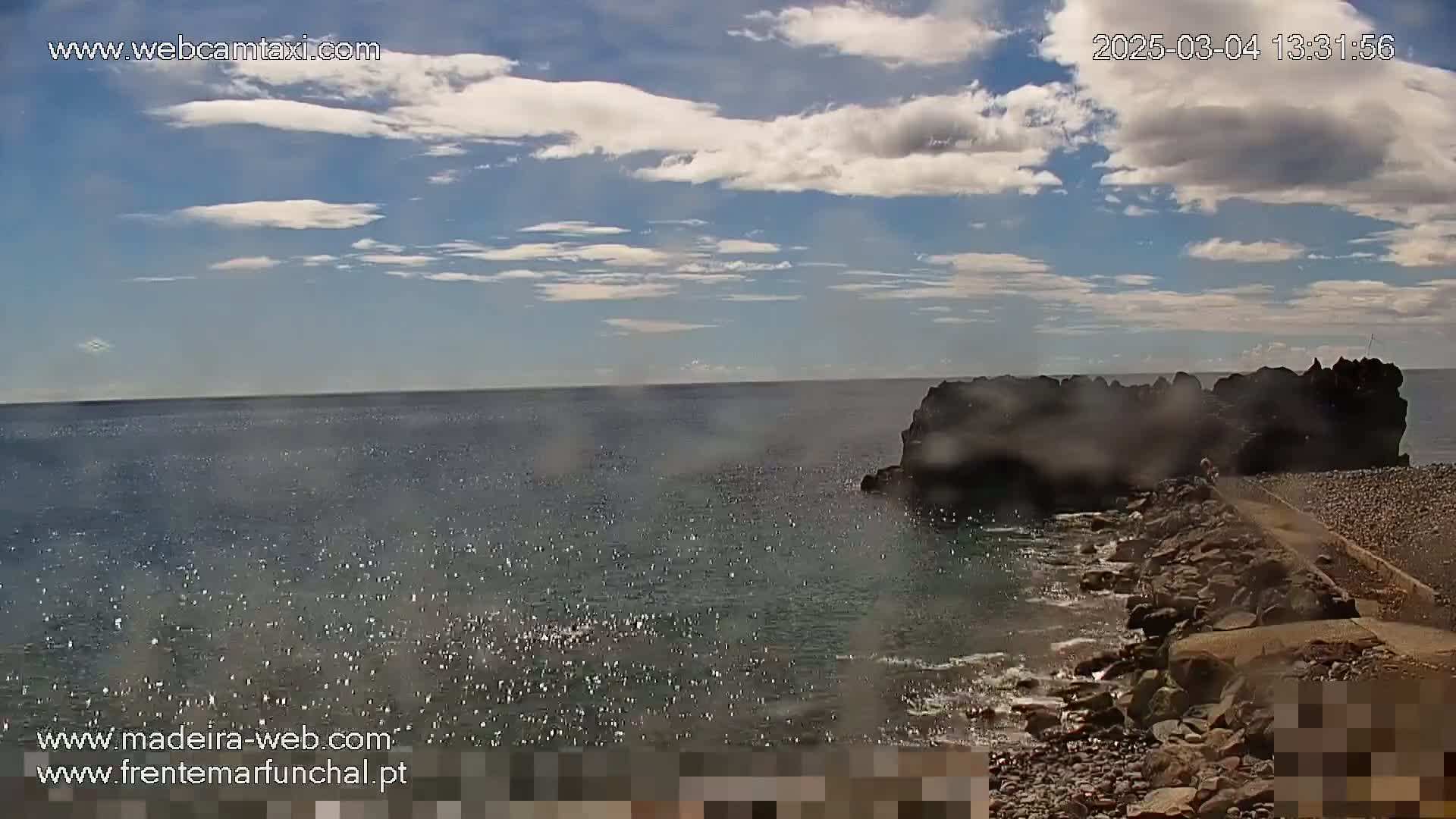 A rocky outcrop juts into a calm, sparkling ocean under a partly cloudy sky.