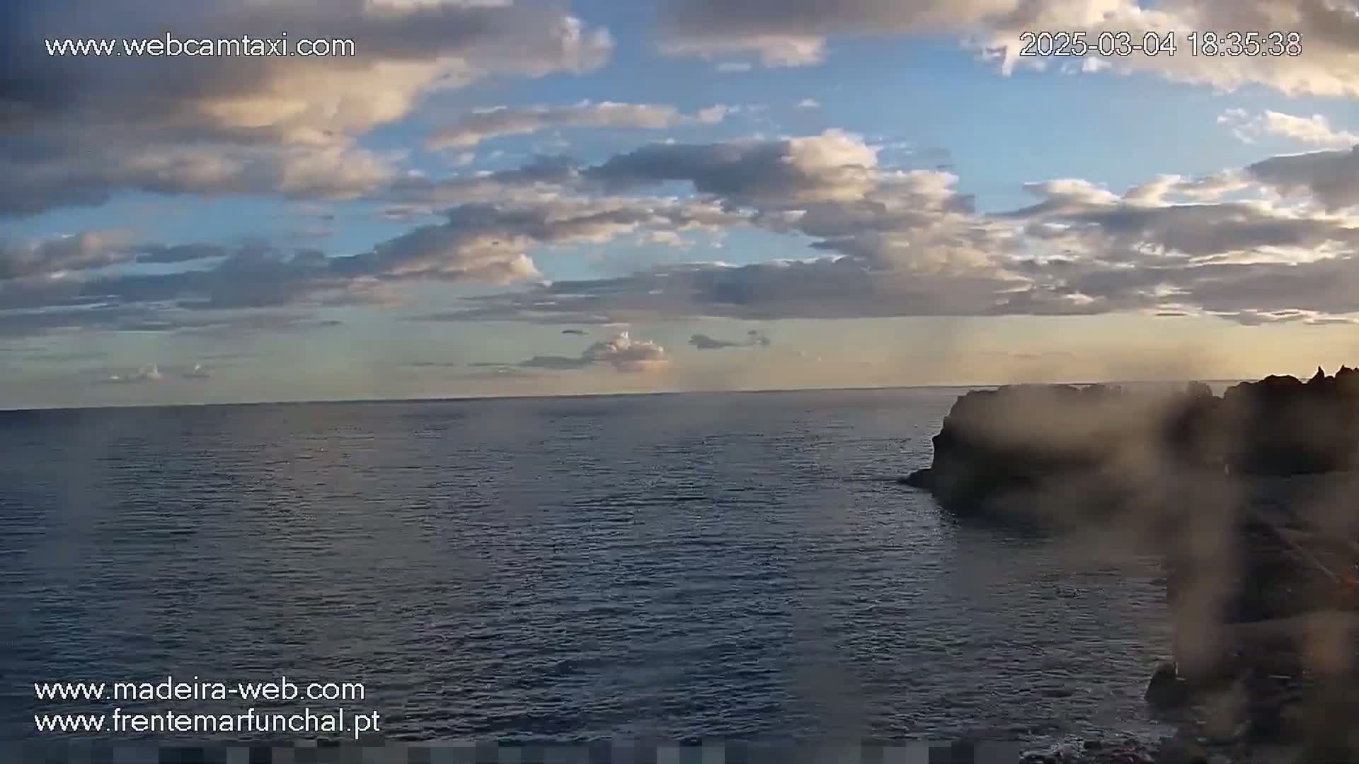 A partly cloudy evening sky over a dark, calm ocean with dark rocks visible in the foreground.