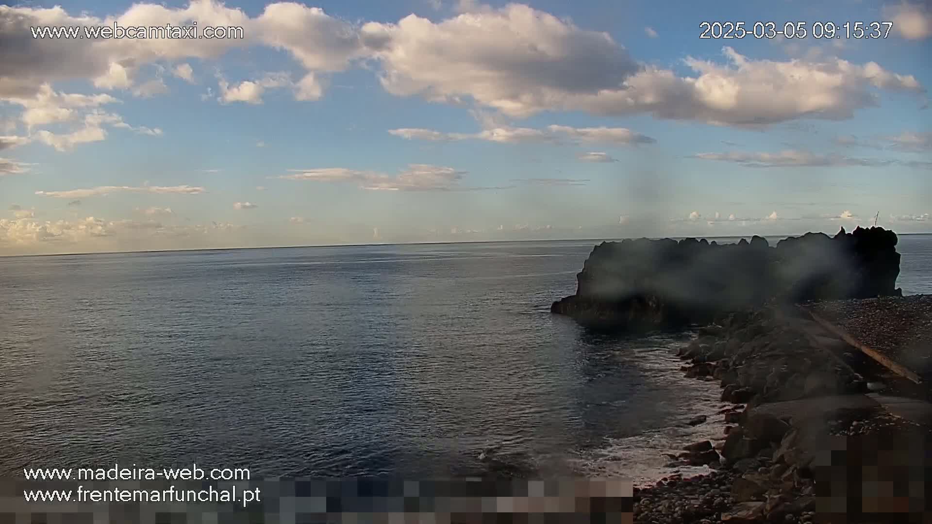 A partly cloudy sky overlooks a dark, rocky shoreline meeting a calm ocean.