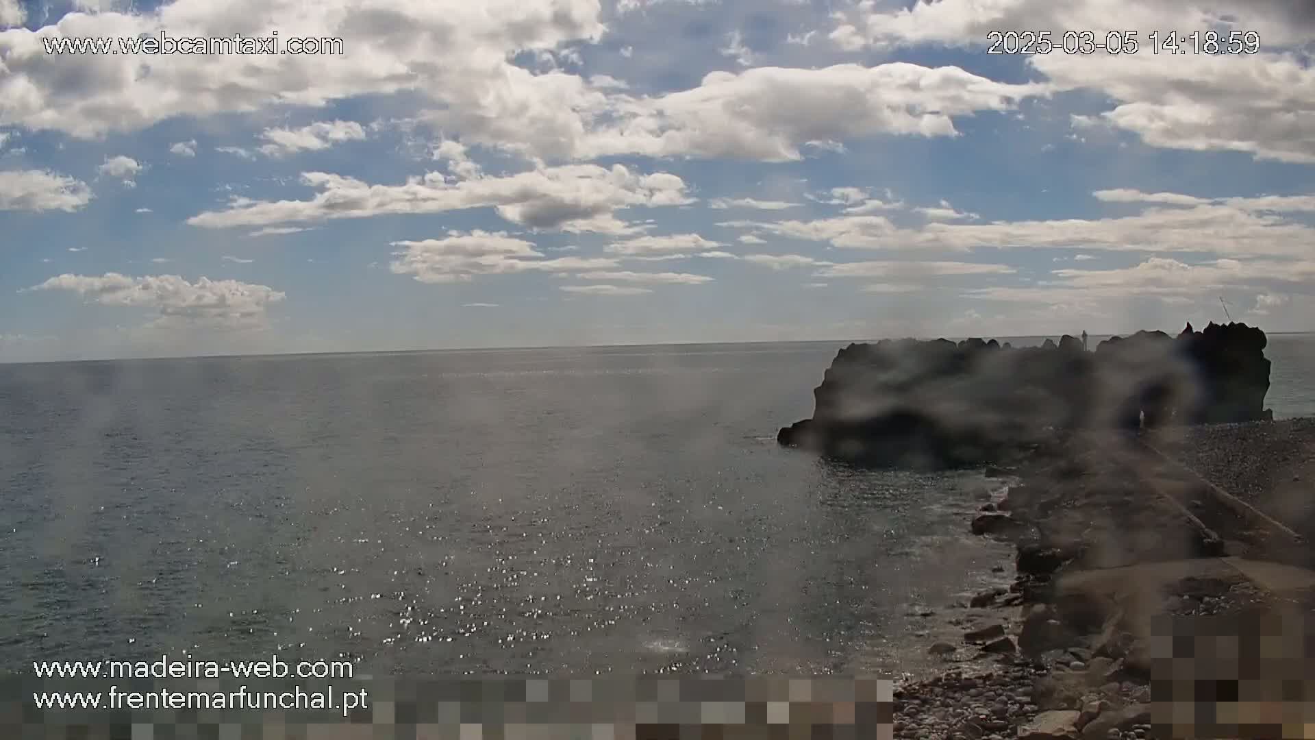 A partly cloudy sky overlooks a dark, rocky shoreline and calm ocean water.