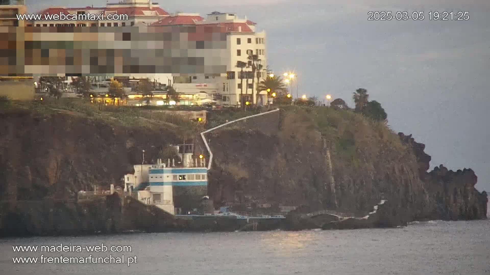 A multi-story building sits atop a dark, rocky cliff overlooking a calm body of water under an overcast sky.