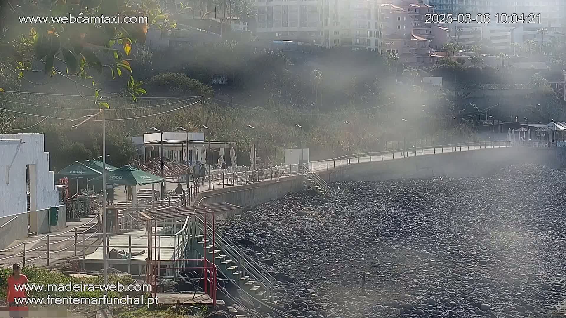 A hazy, sunny day reveals a rocky shoreline with a paved walkway, buildings, and outdoor seating areas along a coast.
