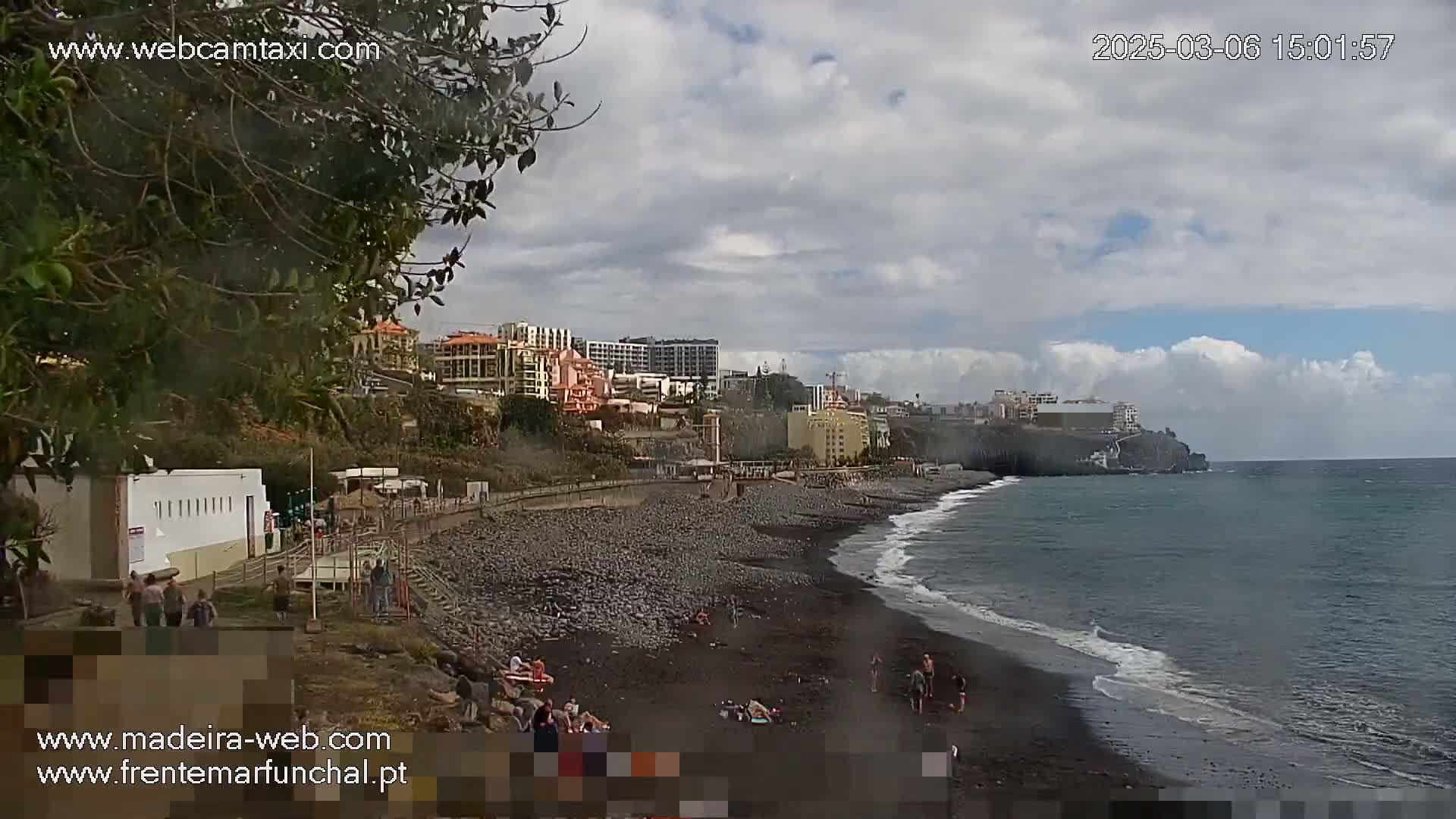 A partly cloudy day shows a black sand beach with people relaxing and buildings along the coast, next to a calm ocean.