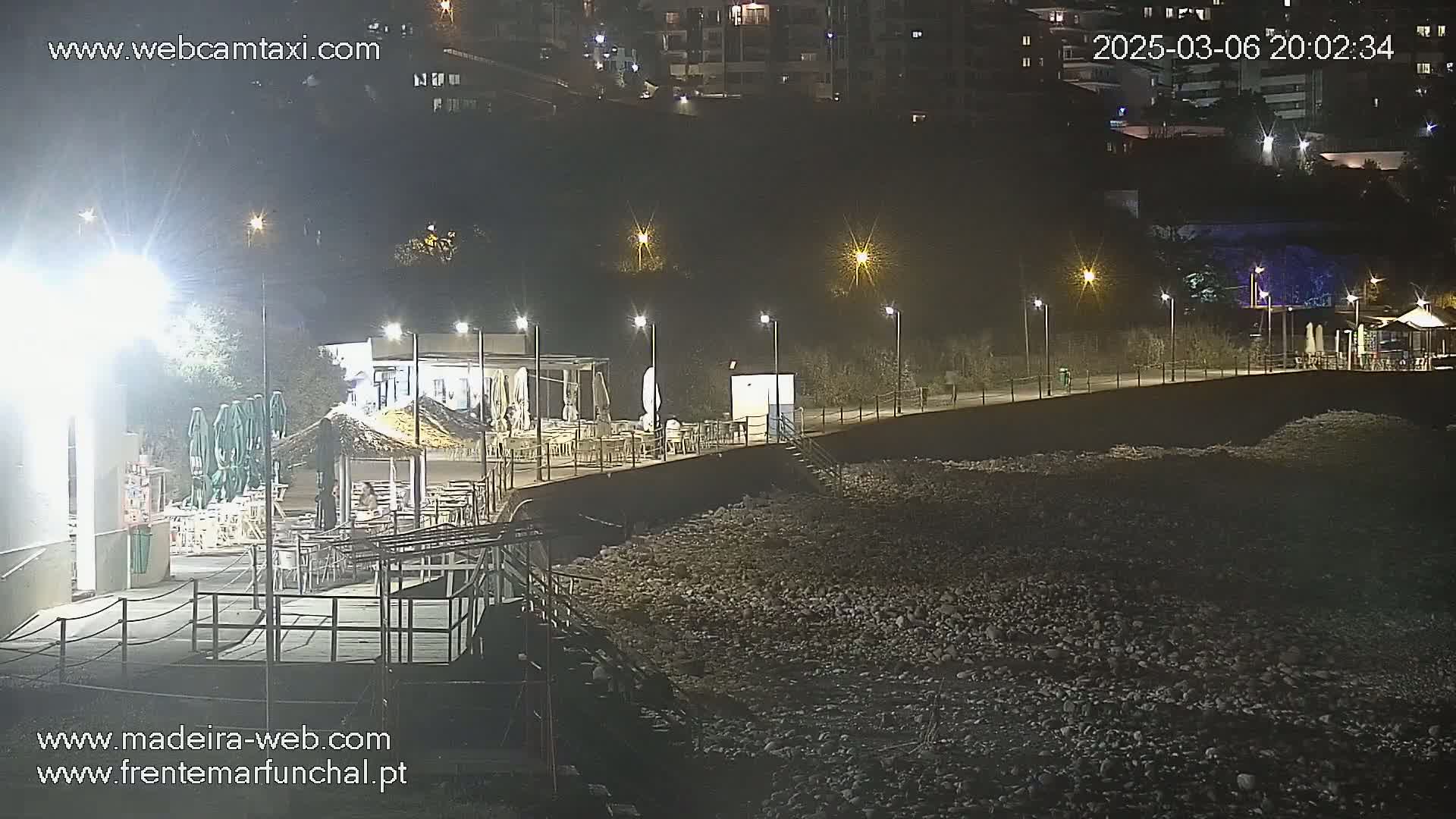 A nighttime view of a rocky beach with an outdoor restaurant and several lights illuminating the scene.