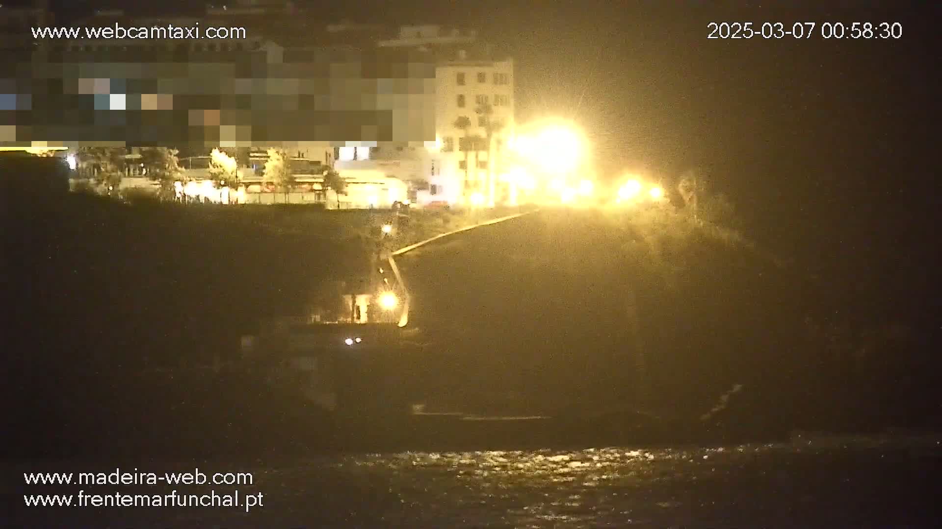 At night, illuminated buildings and a roadway are visible above a dark, rocky shoreline with mildly disturbed water.