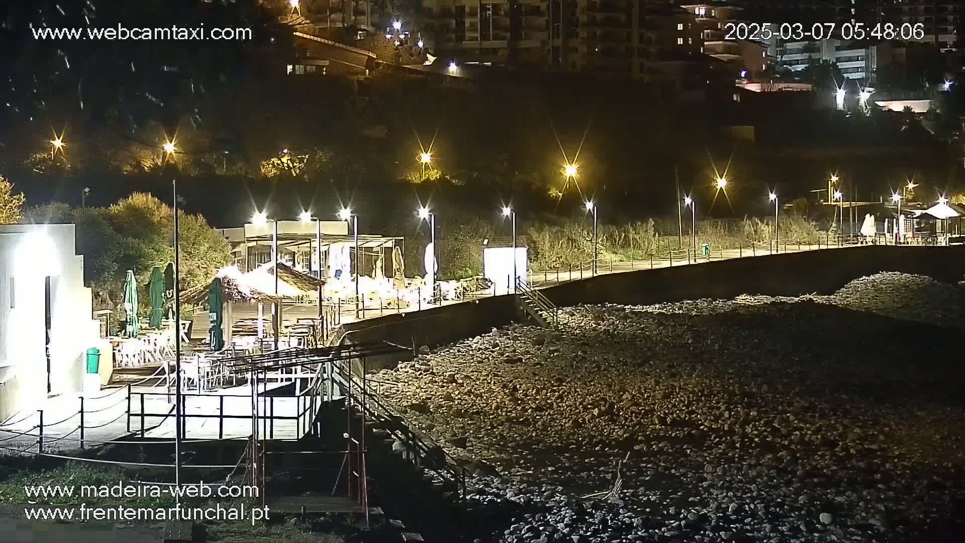 At night, a rocky beach is illuminated by streetlights, with a restaurant and boardwalk visible in the background.