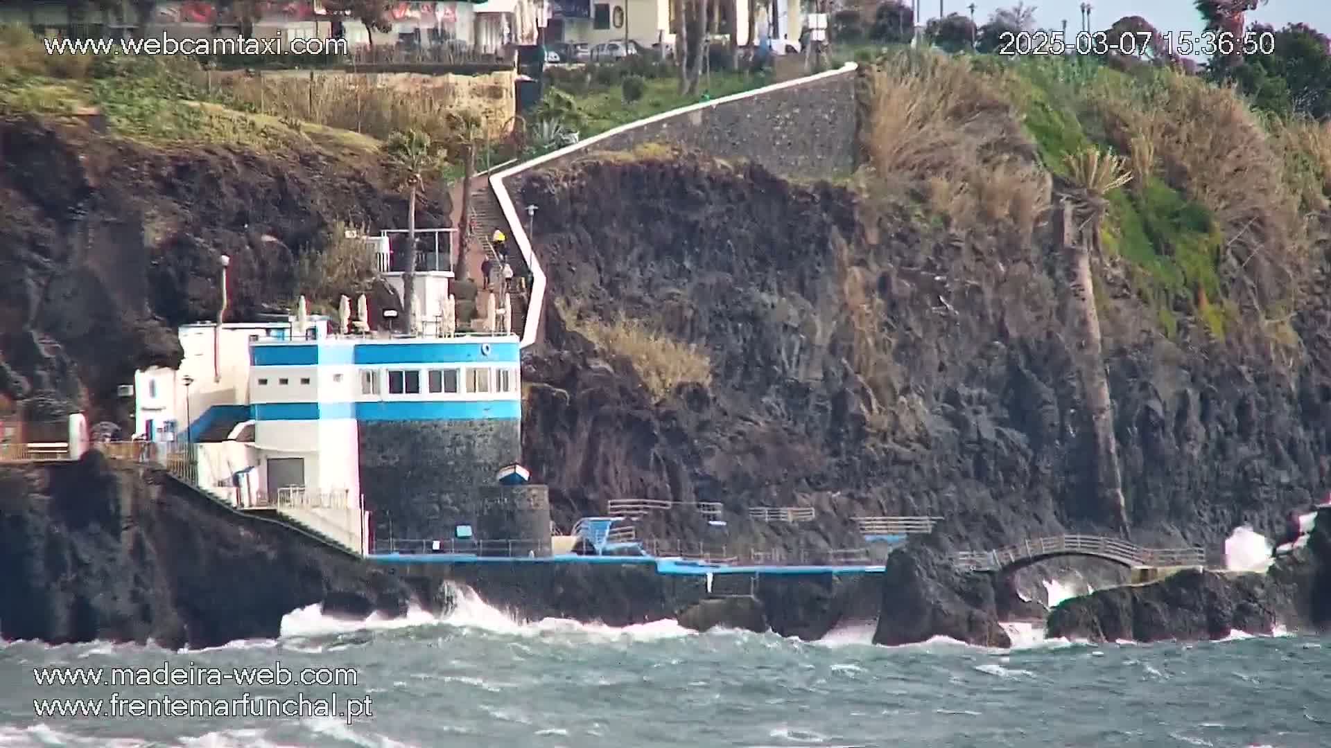 A small, blue-and-white building sits on a rocky cliff overlooking a choppy ocean with waves crashing against the shore.