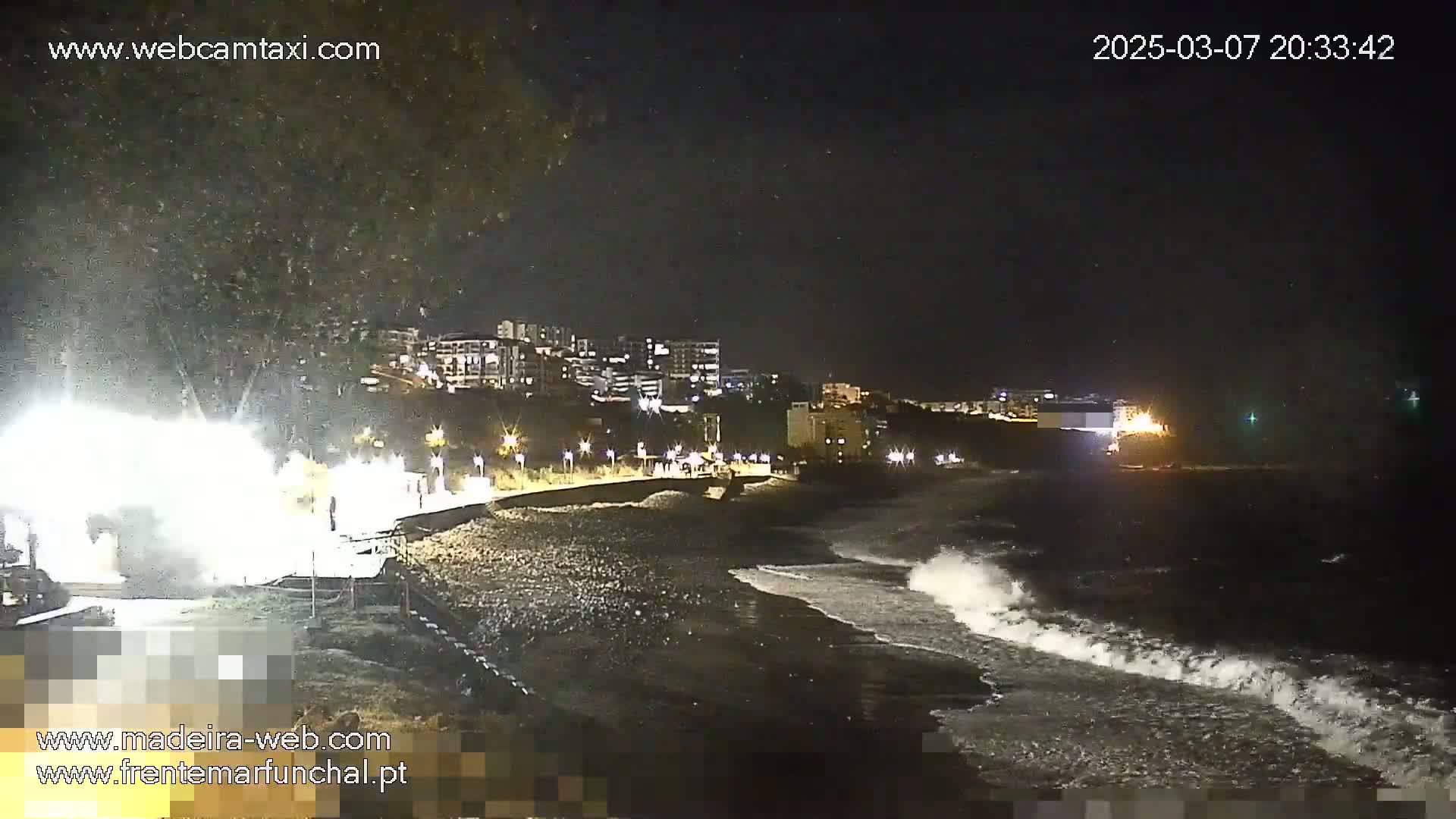At night, under a dark sky, moderately sized waves crash on a dark beach near a city's illuminated shoreline.
