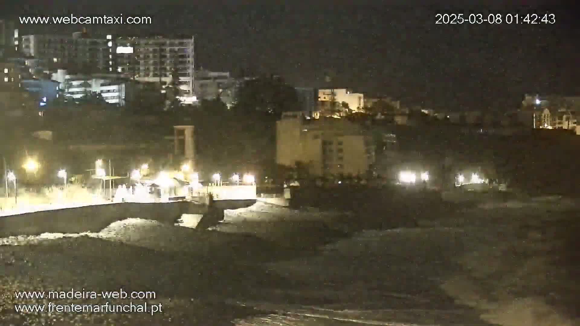 Nighttime view of a dark ocean with waves crashing against a shoreline, illuminated by lights from buildings and streetlights in a city overlooking the water.