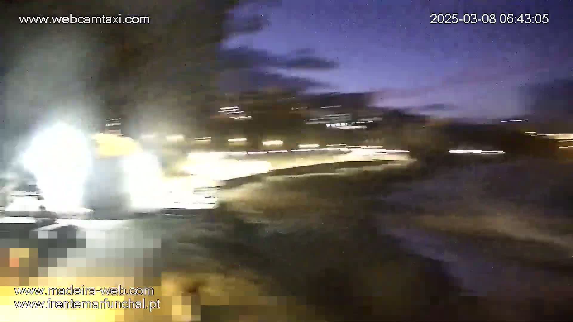 A blurry, nighttime image shows bright lights and white spray from what appears to be a vehicle moving at high speed near a waterfront area under a dark, twilight sky.