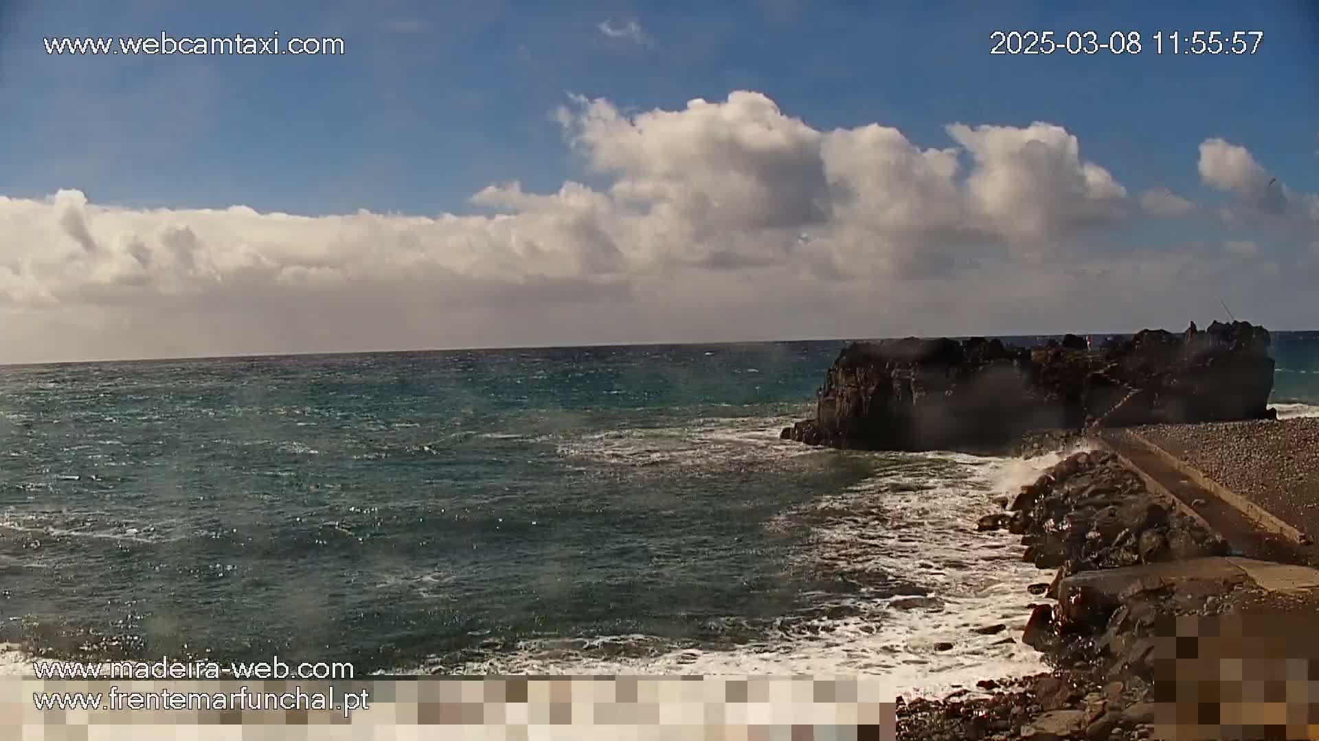 A partly cloudy day shows a rocky outcrop extending into a moderately wavy ocean.