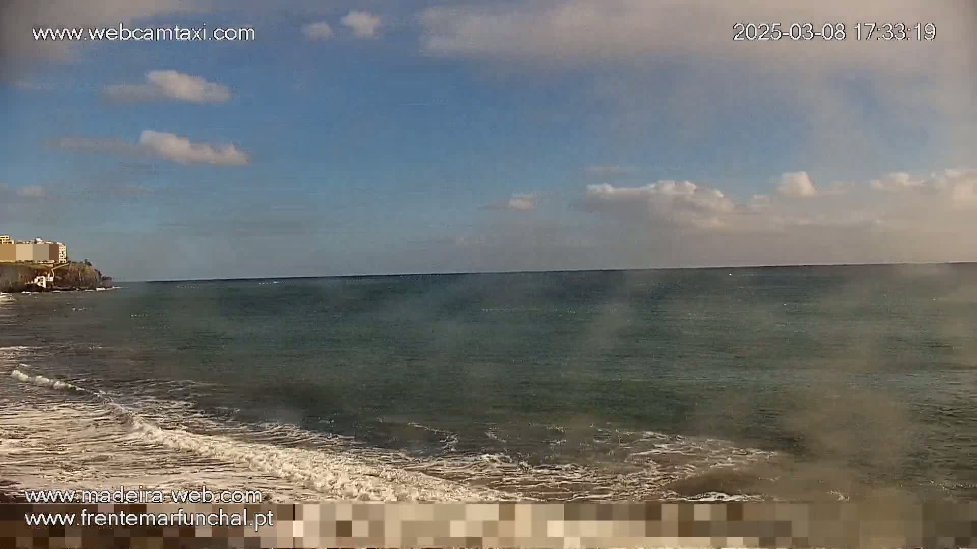 A mostly sunny day shows a calm ocean with small waves lapping a sandy beach near a rocky cliff with buildings on top.