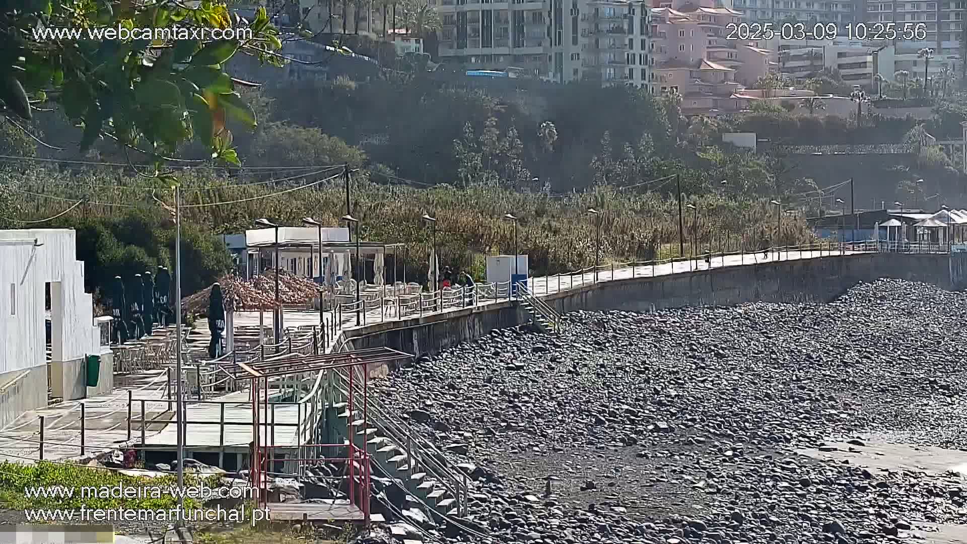 A paved walkway overlooks a rocky beach, with buildings and vegetation visible in the background under a hazy sky.