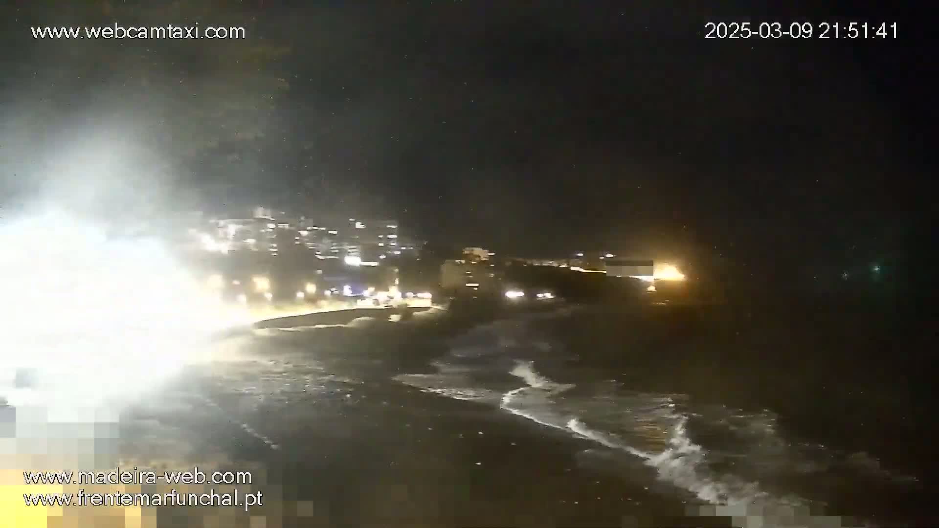 Large waves crash on a dark beach at night near a brightly lit town under a dark, stormy sky.
