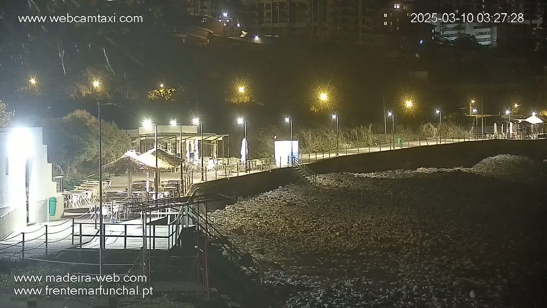 A nighttime view of an outdoor restaurant area along a rocky shoreline, illuminated by artificial lights.