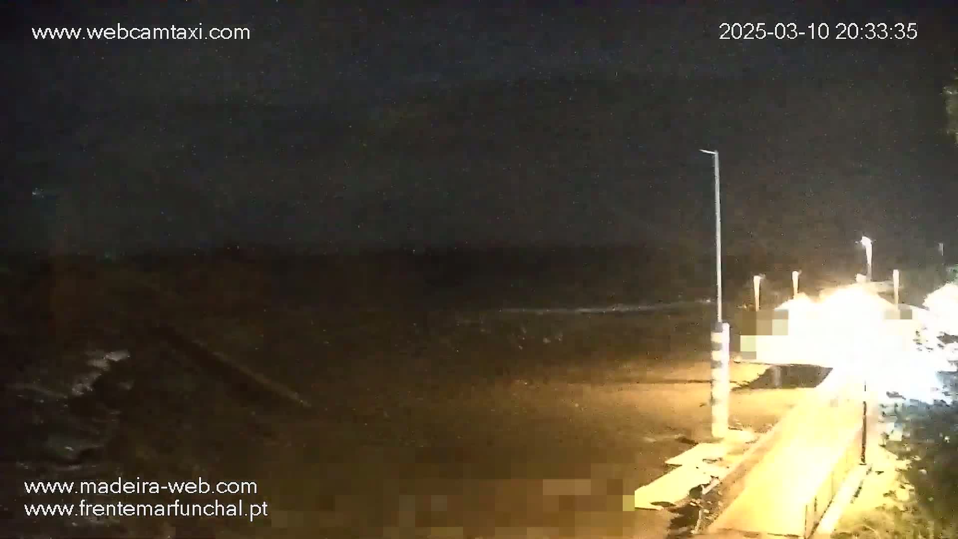A nighttime view of a dark beach with a brightly lit walkway and some vegetation visible to the right, under a clear, starry sky.